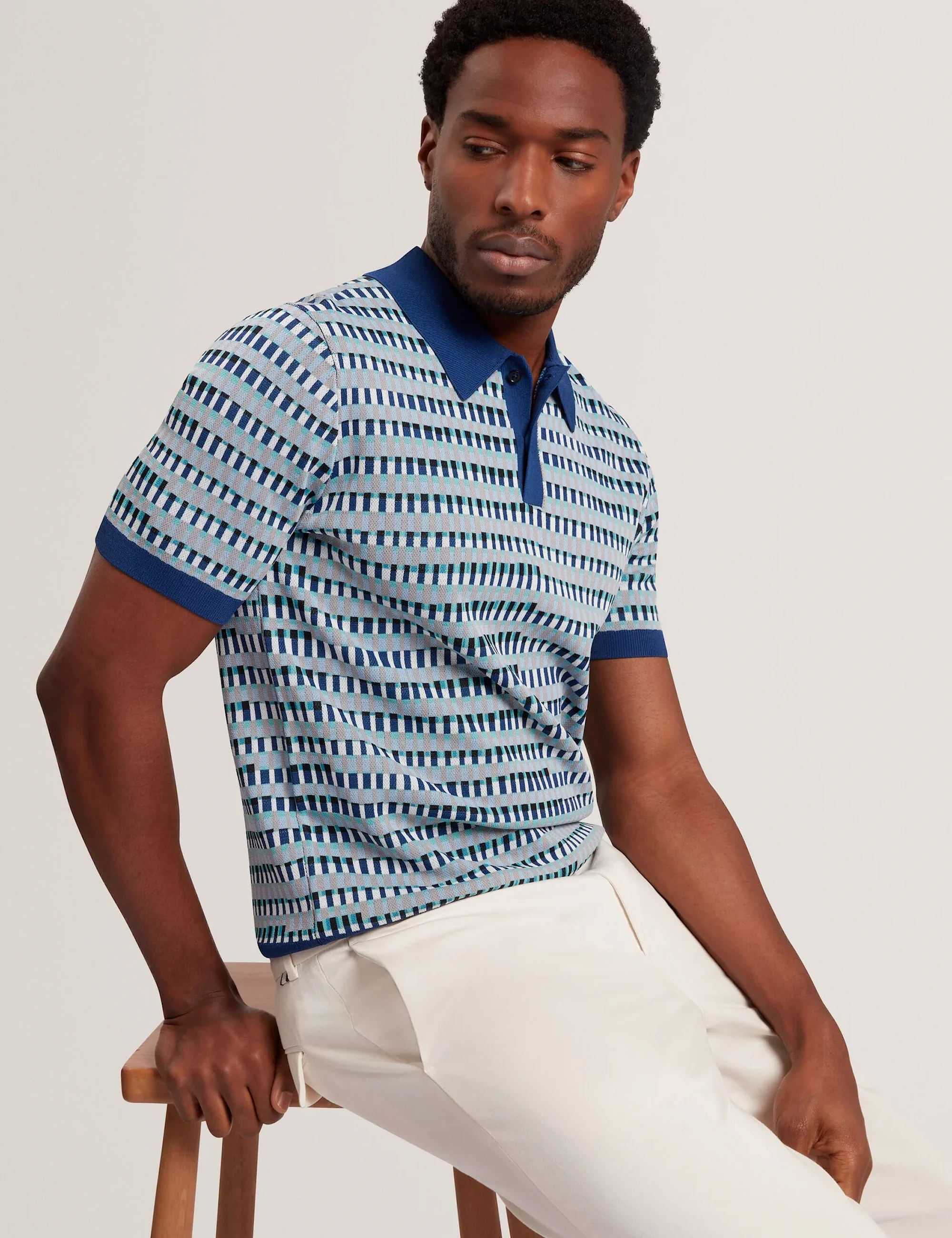 Man in a blue patterned Ted Baker  short-sleeve polo shirt and white pants sitting on a wooden stool.