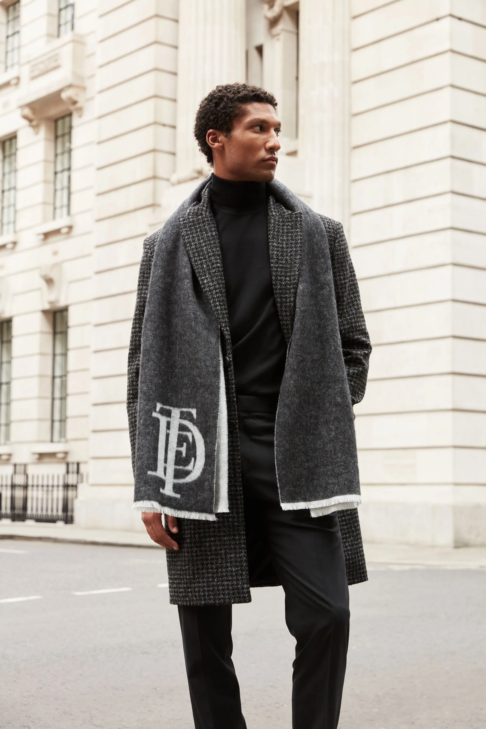 Man wearing a black Ted Baker  turtleneck, dark checkered coat, and a gray scarf with white initials stands on a city street.