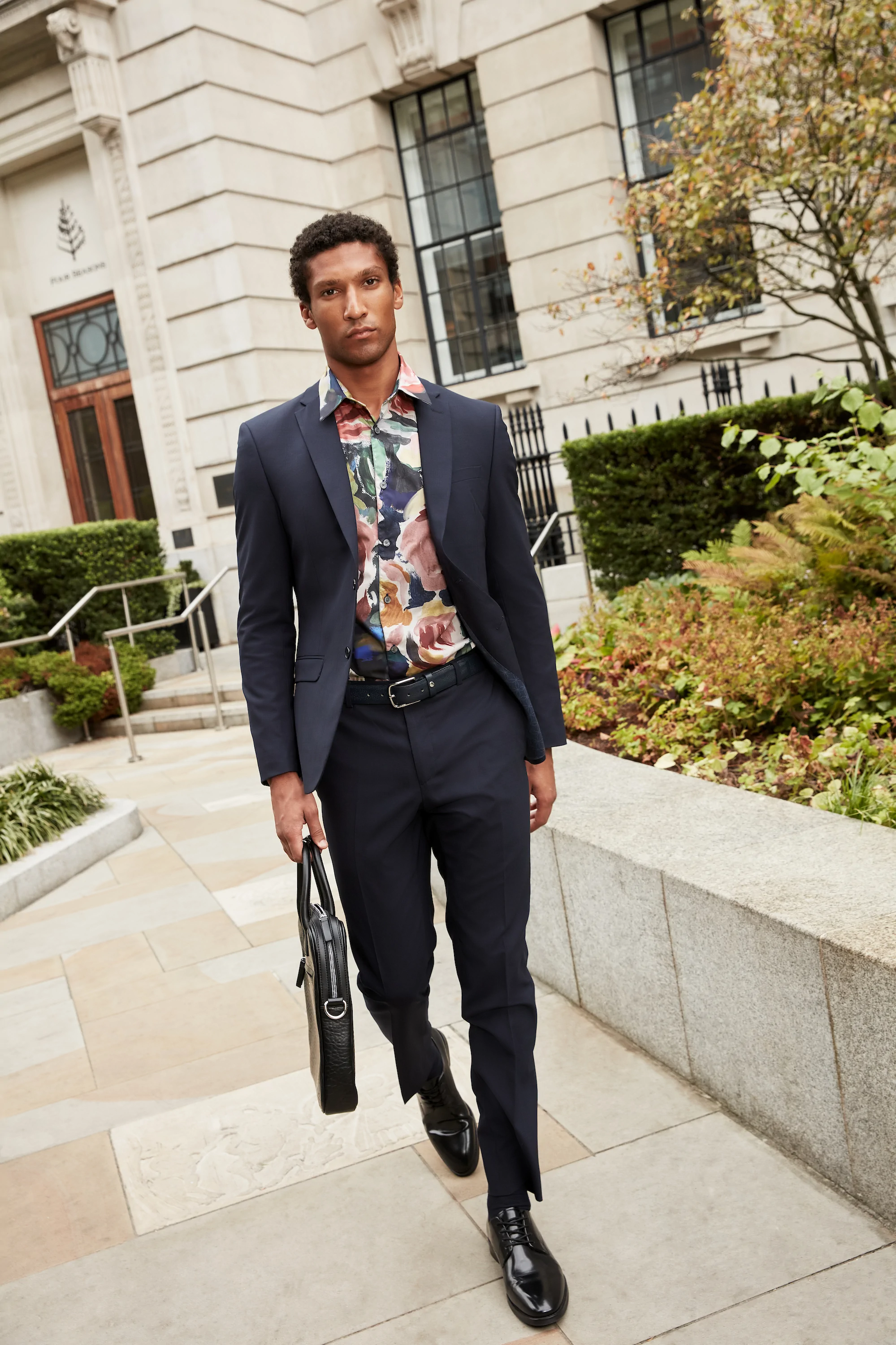 Man walking outdoors wearing a navy  Ted Baker suit with a colorful floral shirt and carrying a black briefcase.