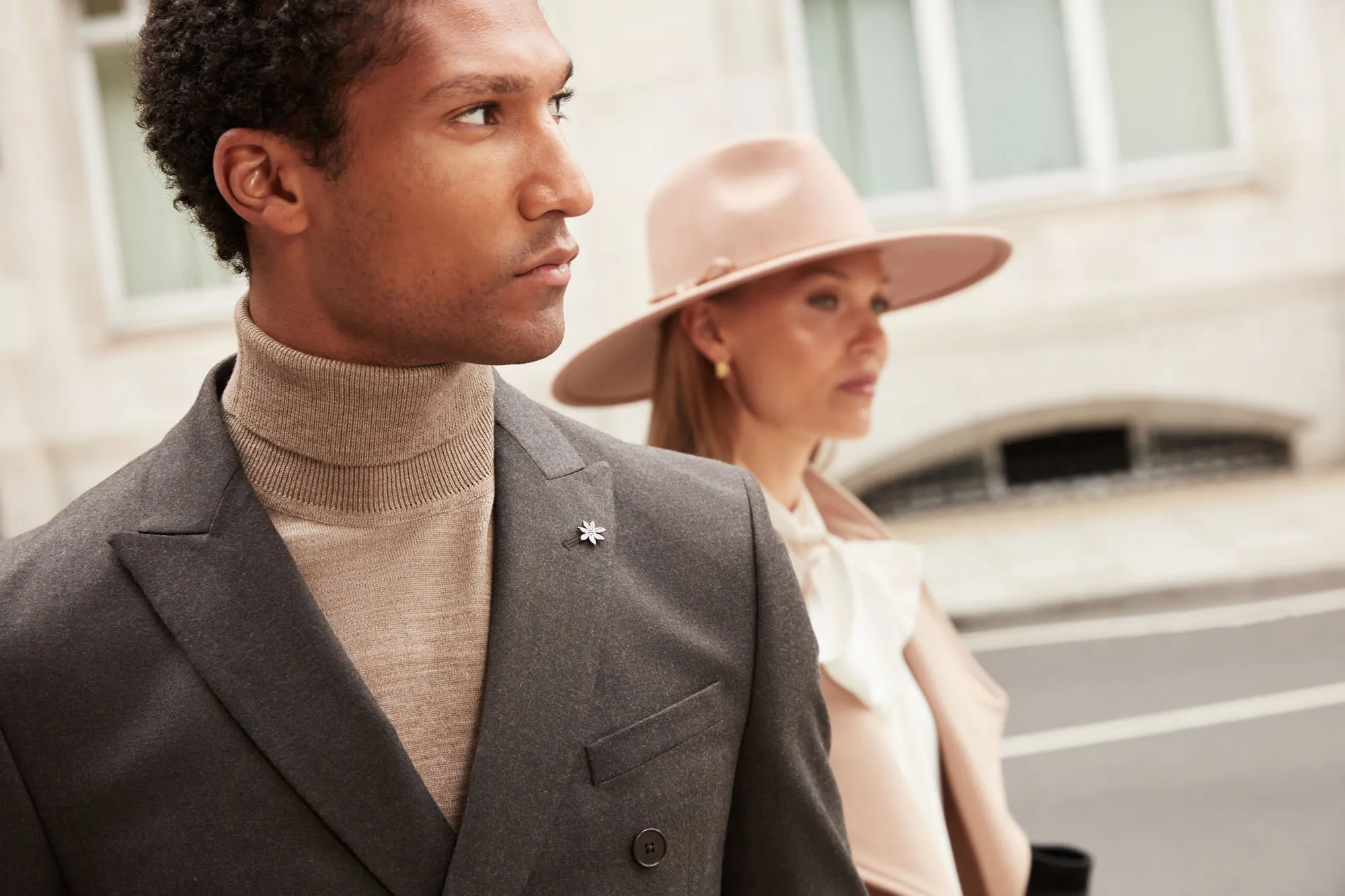 Man in gray Ted Baker blazer and beige turtleneck with woman in beige coat and wide-brimmed hat behind him.