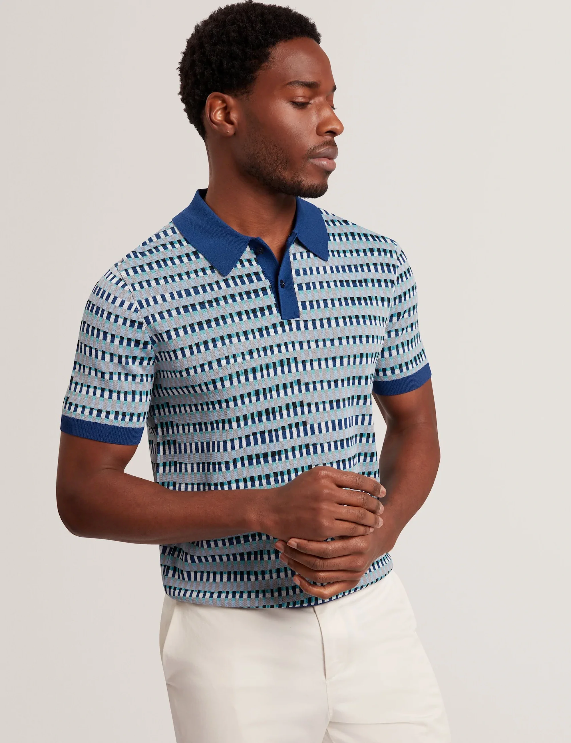 Man wearing a patterned Ted Baker  blue polo shirt with short sleeves and white pants against a neutral background.