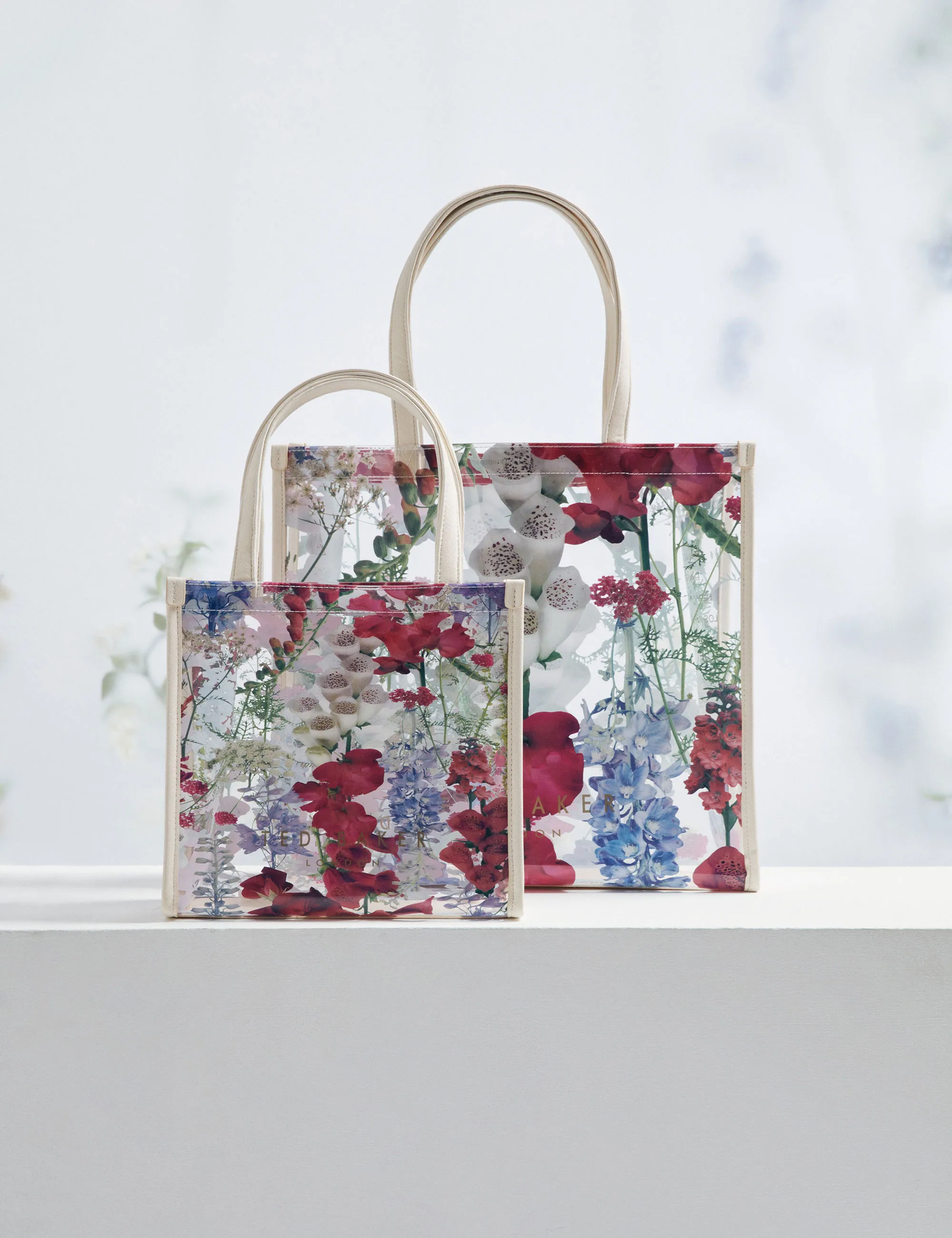 Two transparent Ted Baker tote bags with colorful floral prints and beige handles on a white surface.
