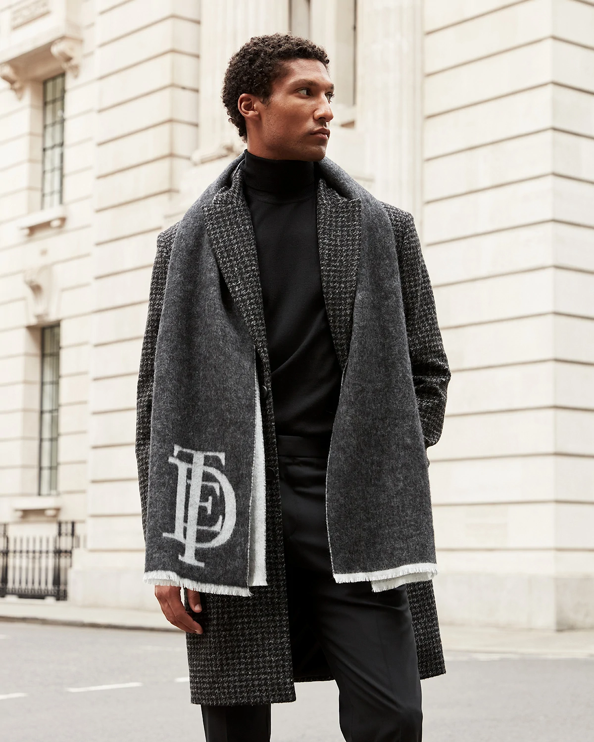 Man wearing a grey Ted Baker houndstooth coat and dark scarf with white logo standing on a city street.