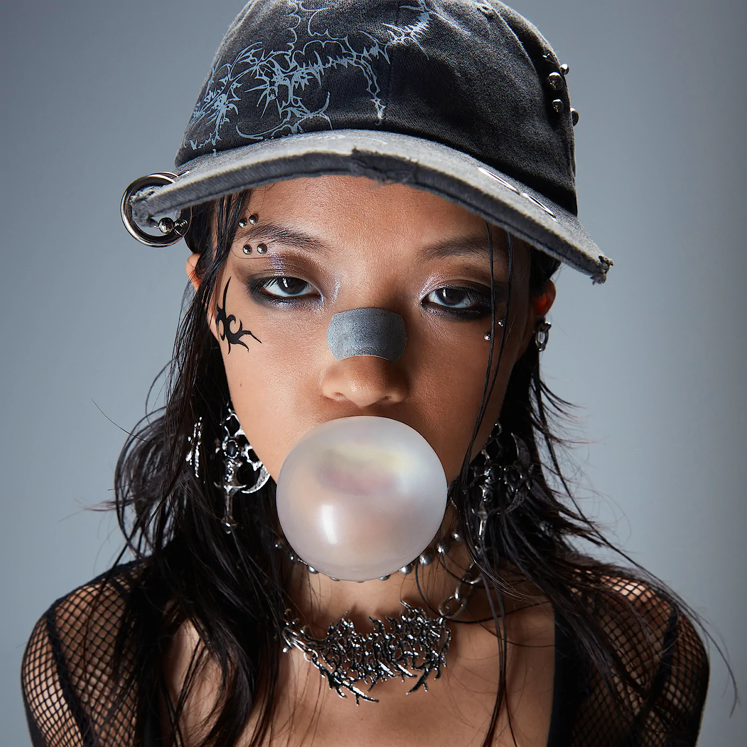 Young woman wearing a black cap, mesh top, silver jewelry, with a nose strip and blowing a bubblegum bubble.