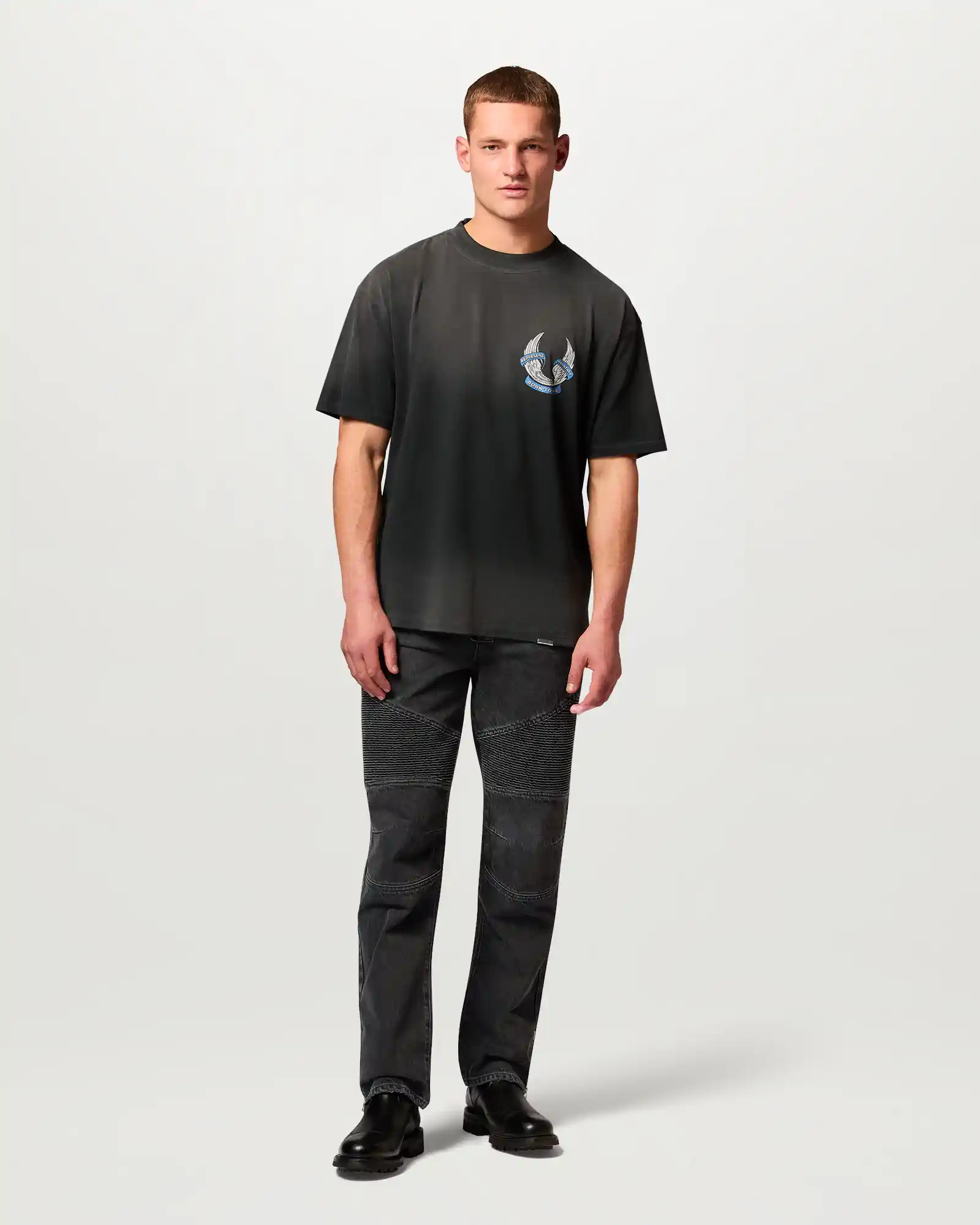 Man wearing a faded black Belstaff t-shirt with a small graphic on the chest and black textured jeans with boots.