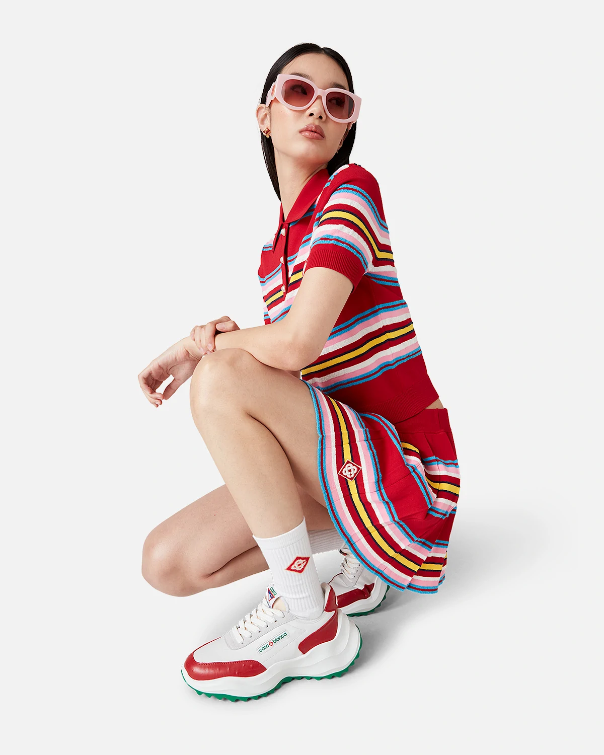 Woman wearing red striped Casablanca polo shirt and skirt with white sneakers and pink sunglasses crouching.
