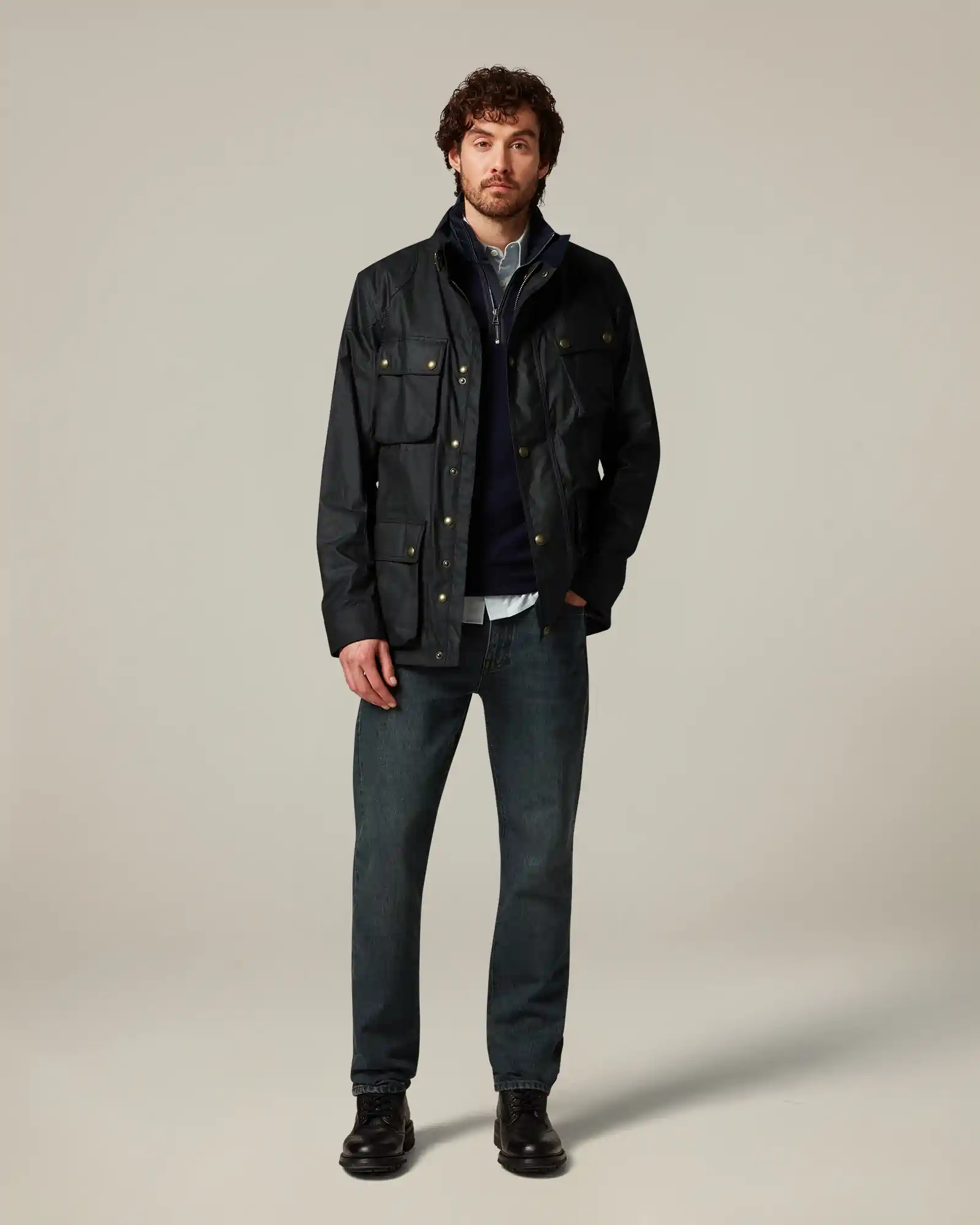 Man standing wearing a dark navy Belstaff jacket with multiple pockets, jeans, and black boots.