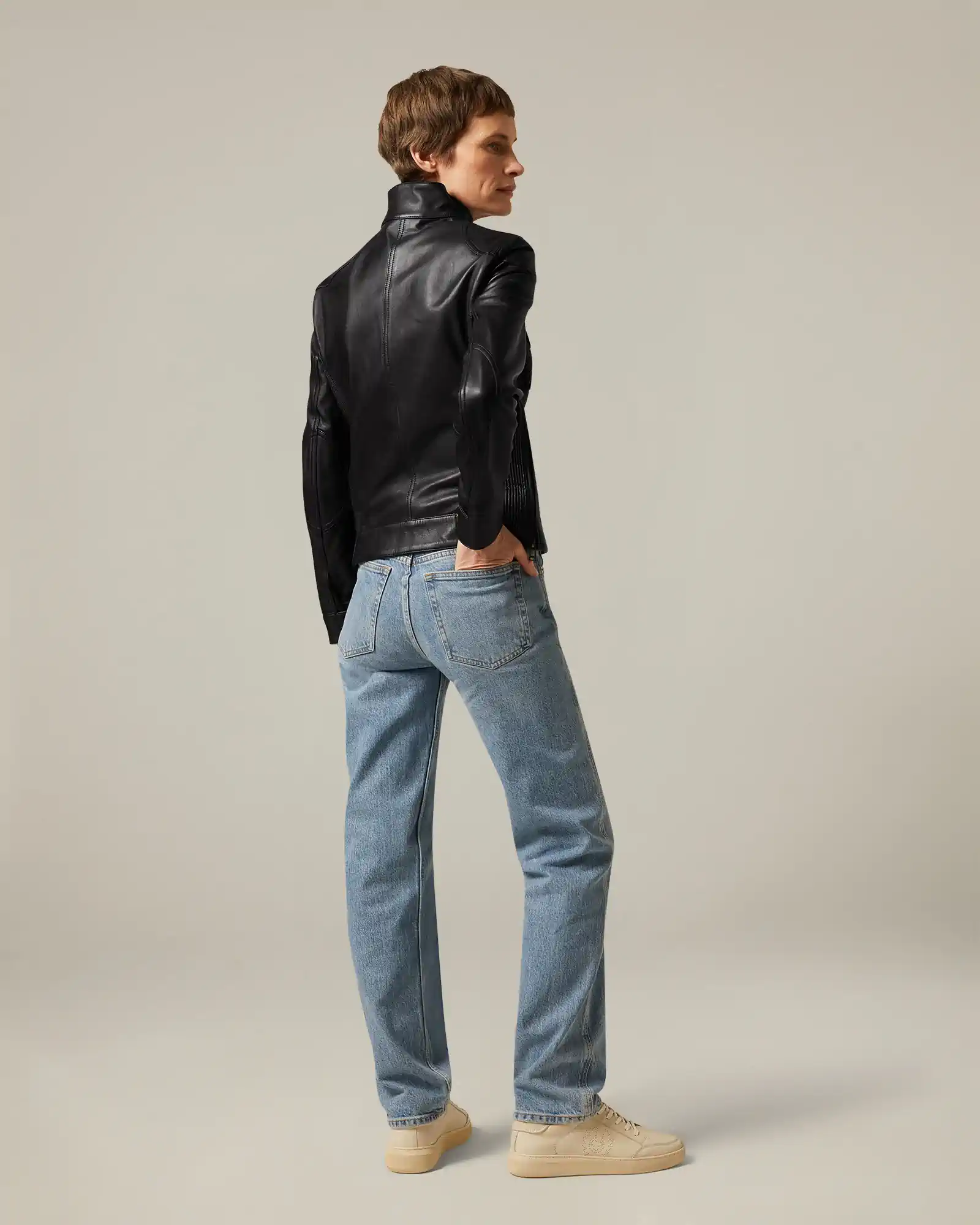 Person wearing a black Belstaff leather jacket, light blue jeans, and beige sneakers facing sideways.
