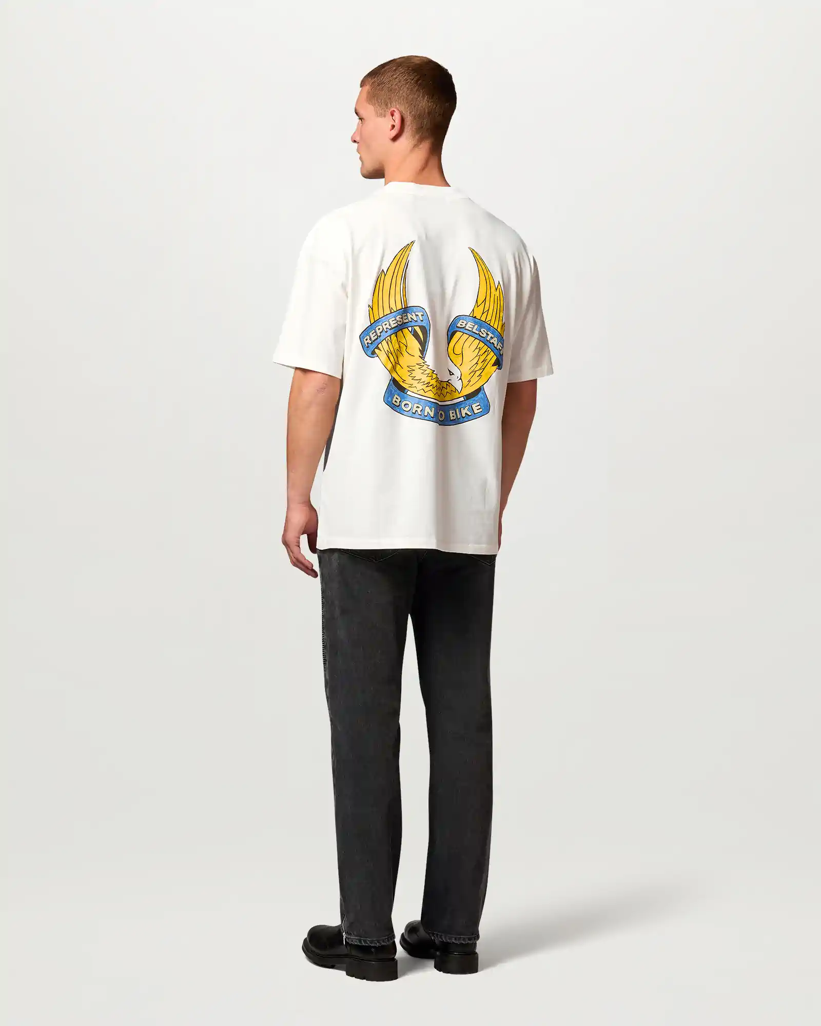 Man wearing white Belstaff T-shirt with yellow eagle and blue text reading Represent Belstaff Born to Bike on back.