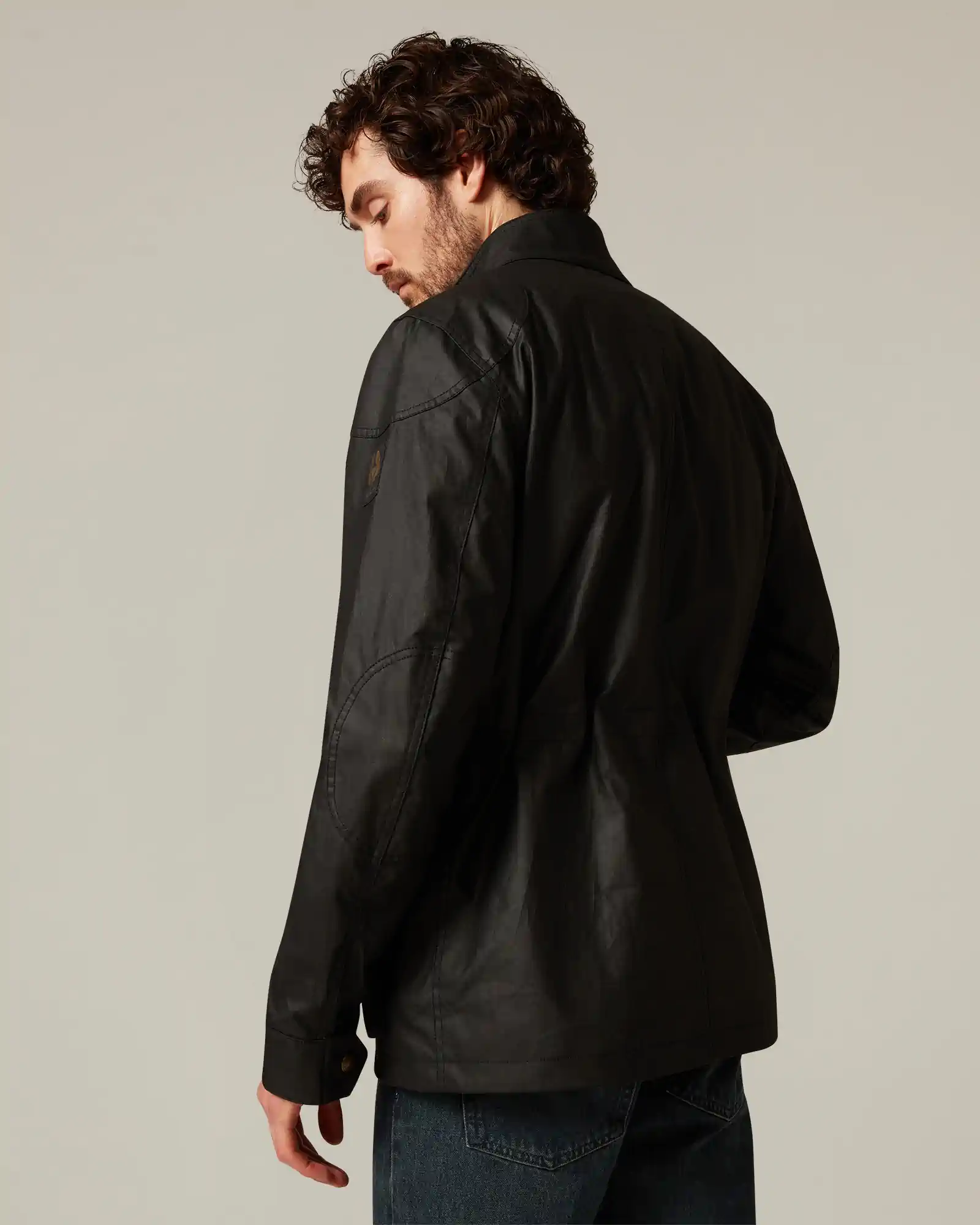 Man with curly hair wearing a black Belstaff jacket viewed from the back against a neutral background.