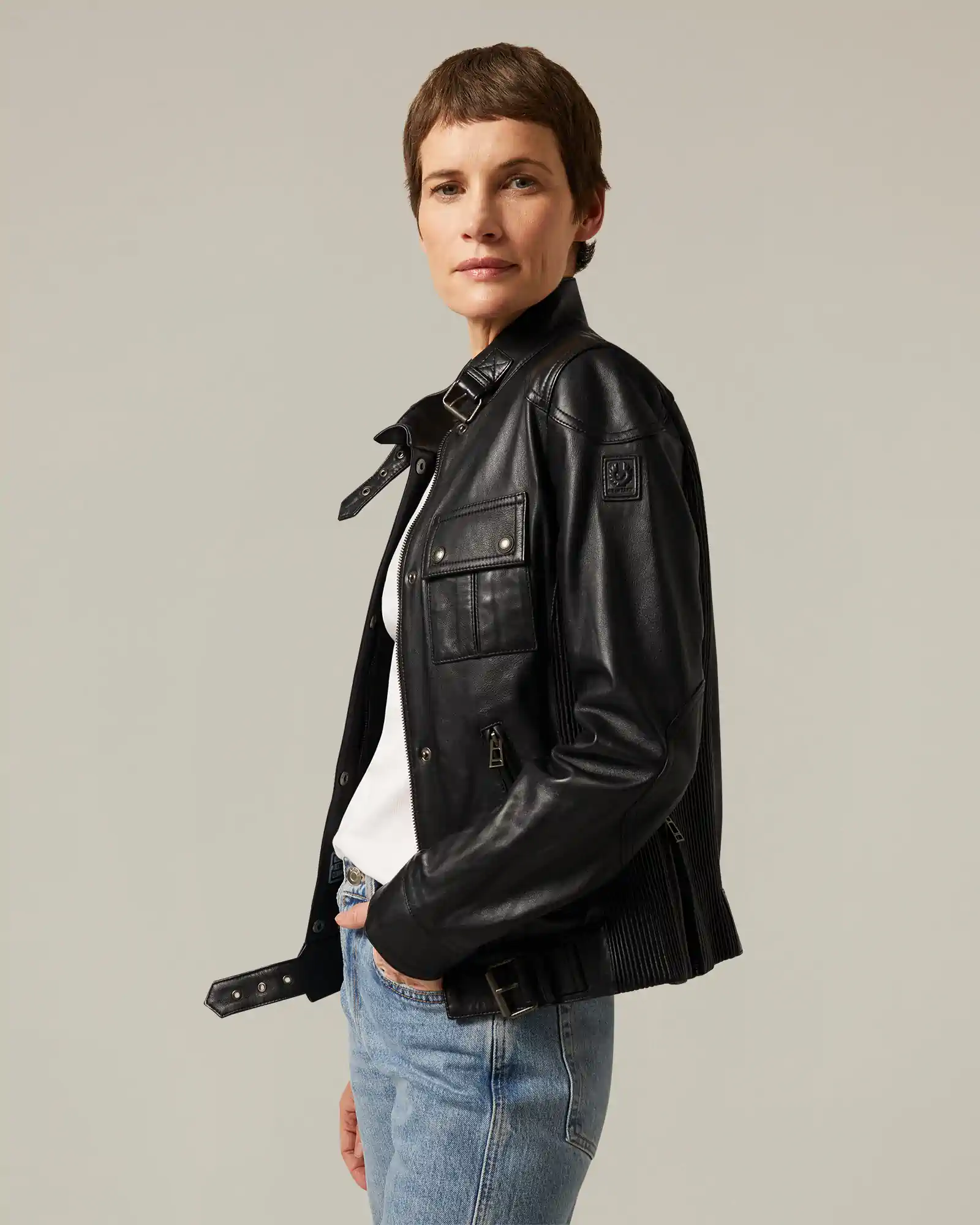 Woman wearing a black Belstaff leather jacket over a white top and blue jeans against a plain background.