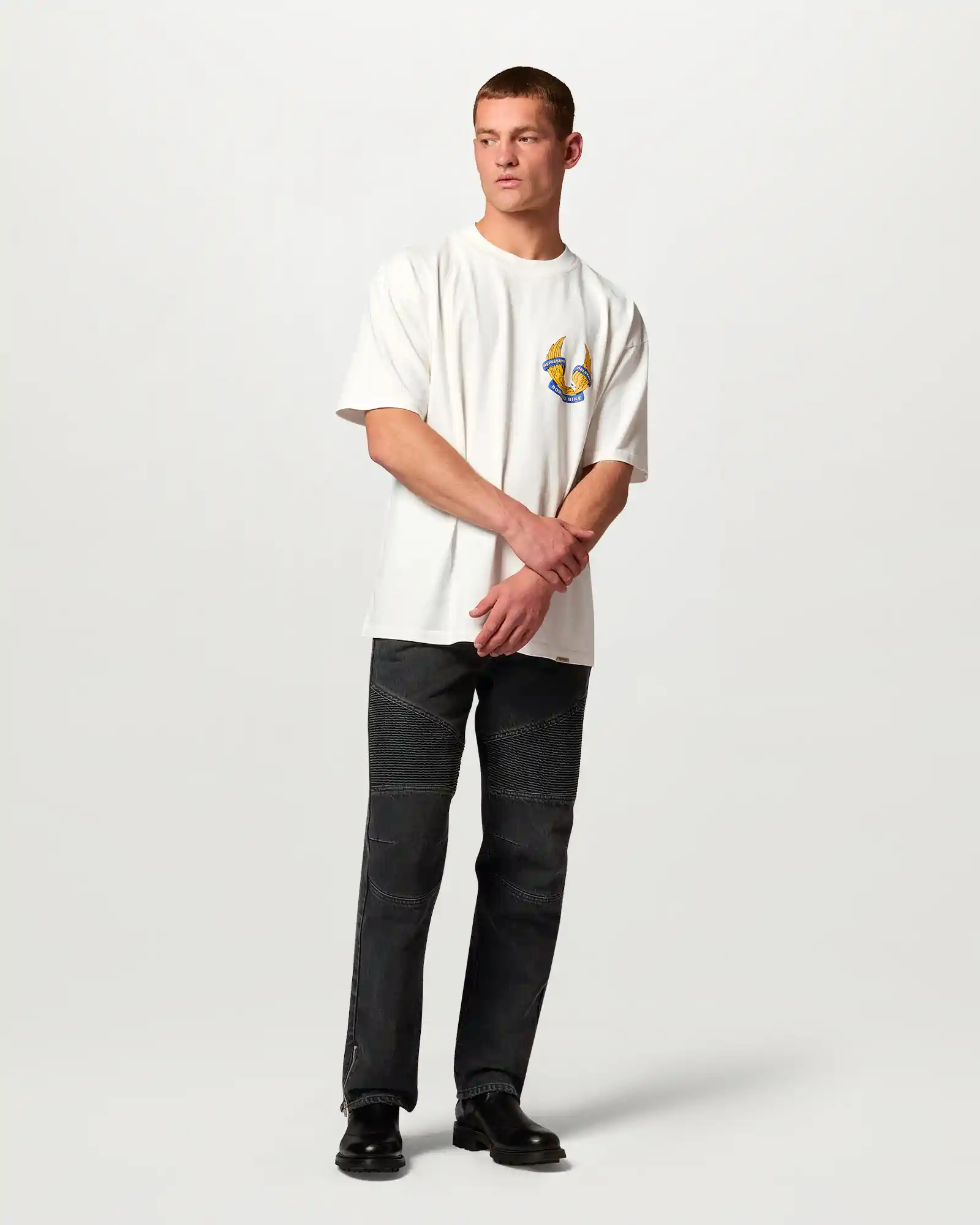 Man wearing white Belstaff t-shirt with bird logo and black pants with ribbed knee design and black shoes.