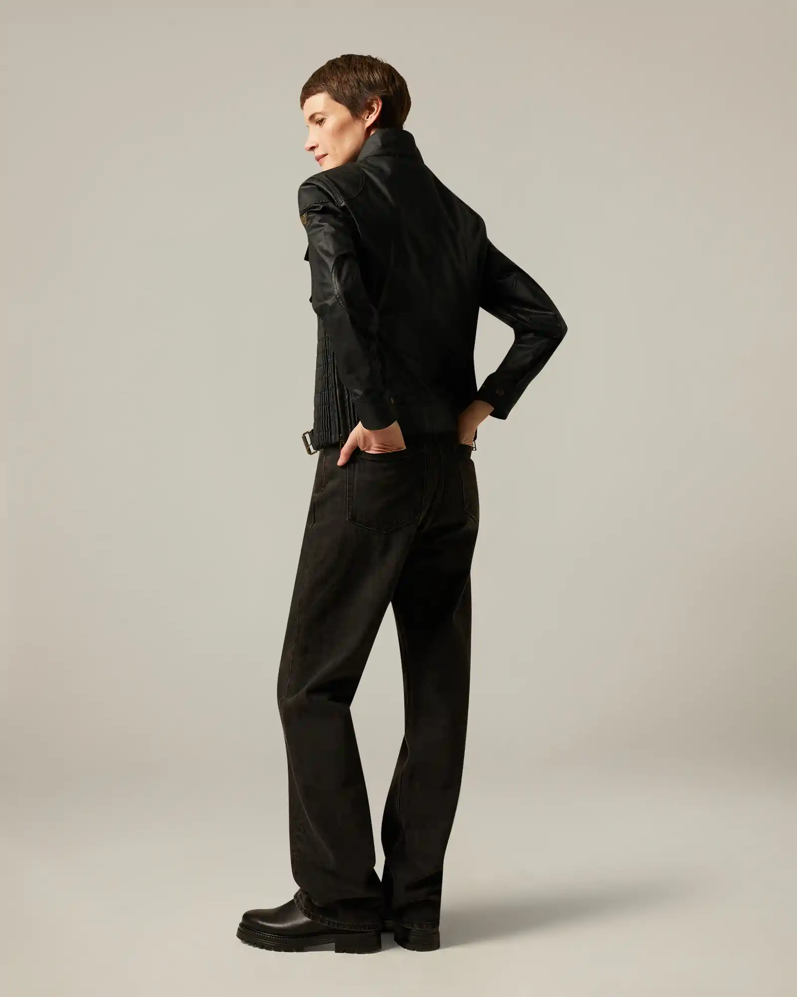 Back view of a woman wearing a black Belstaff leather jacket, black pants, and black boots on a plain background.