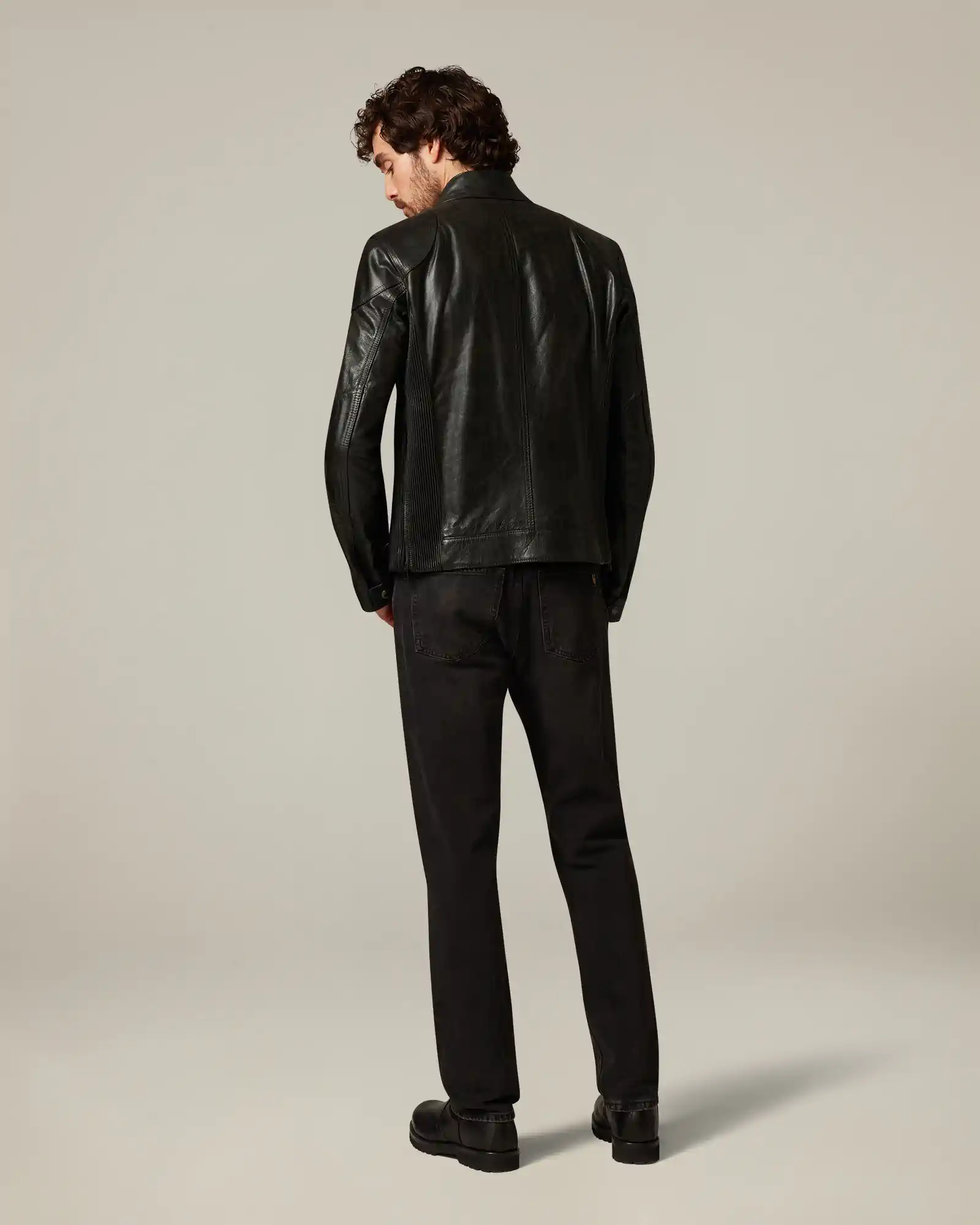 Man wearing black Belstaff leather jacket and black pants, facing away against a plain background.