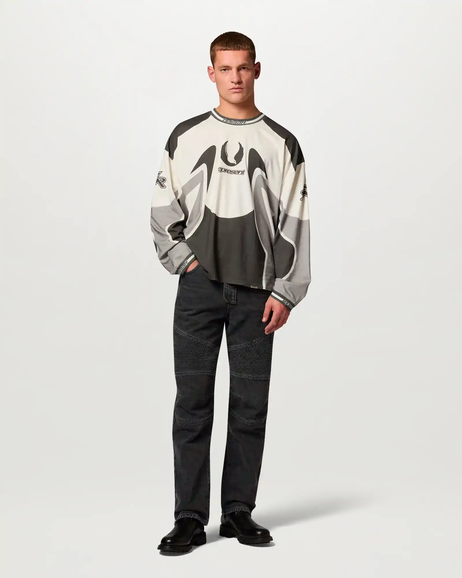Man wearing a graphic Belstaff long-sleeve shirt and black pants with textured knee panels and black boots.