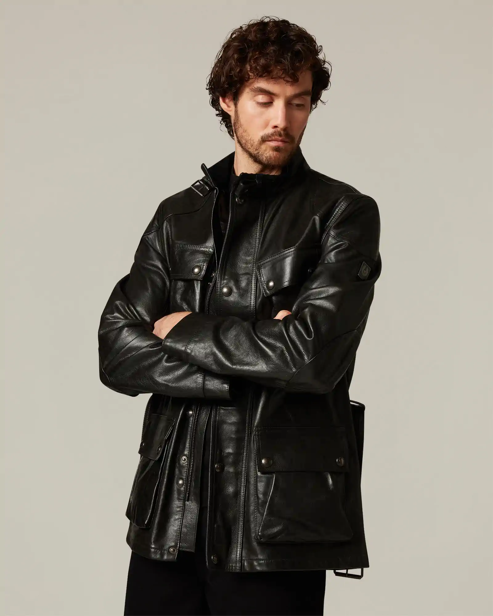 Man wearing a black Belstaff leather jacket with multiple flap pockets and a high collar, arms crossed.