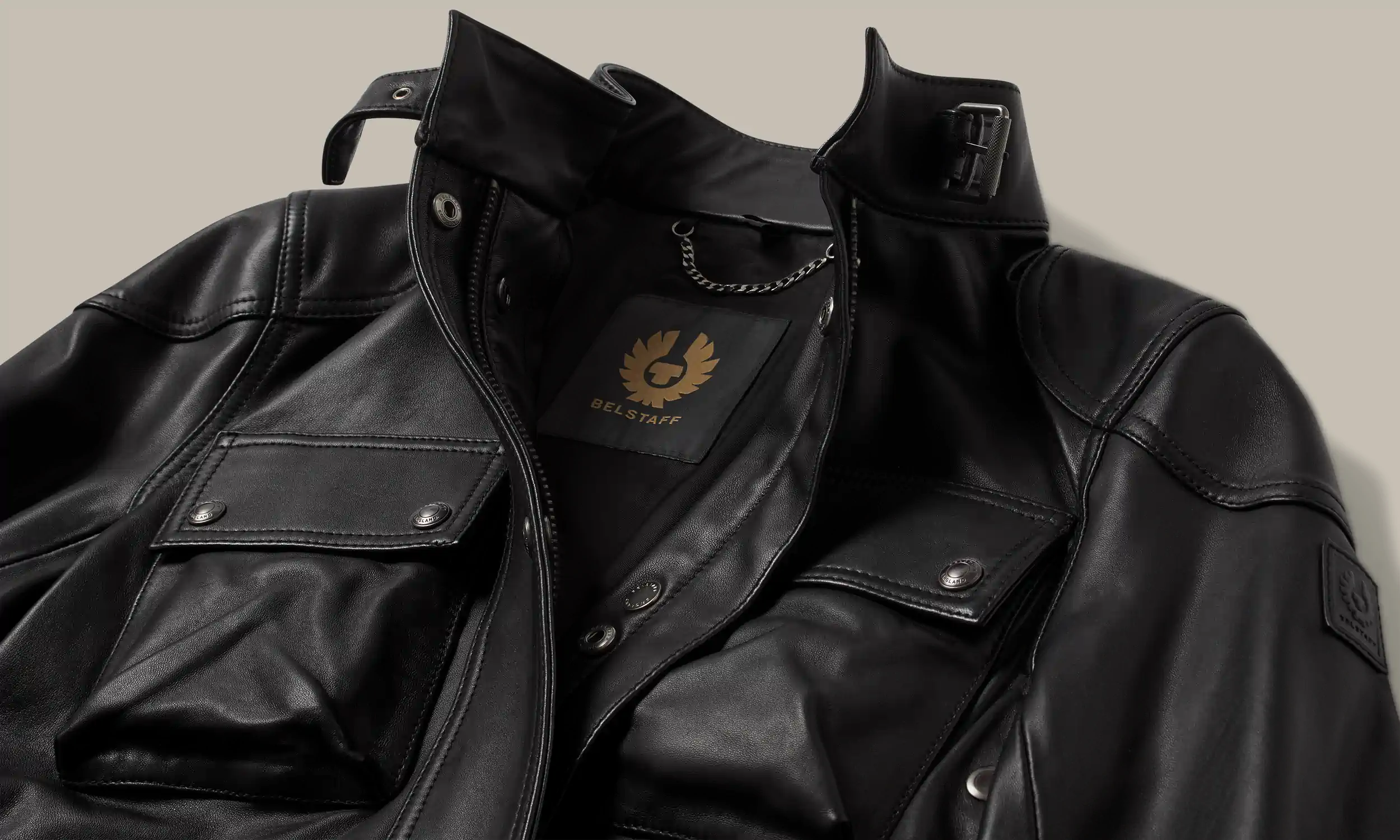 Close-up of a black Belstaff leather jacket showing pockets, collar buckle, and logo patch on sleeve.