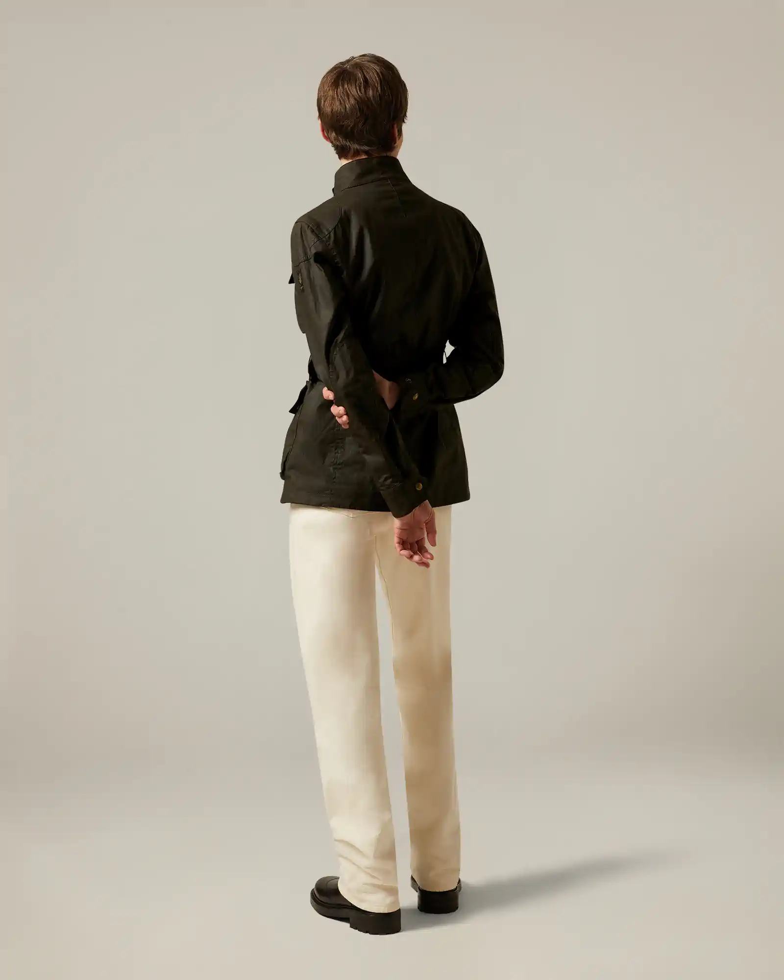 Back view of a person wearing a dark Belstaff jacket, cream pants, and black shoes standing against a plain backdrop.