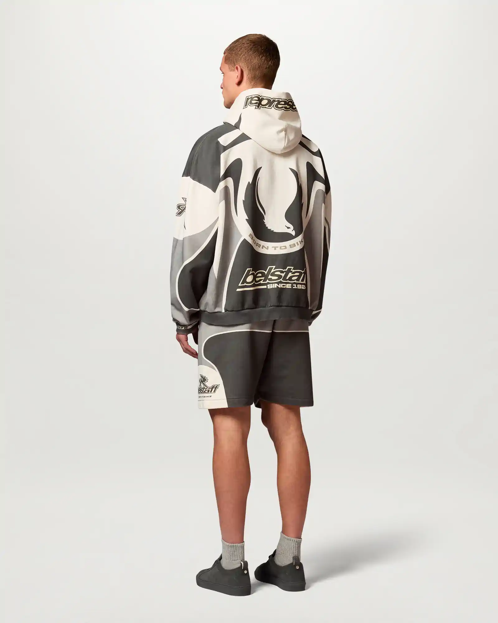 Man seen from back wearing a black and white Belstaff graphic hoodie with matching shorts and gray sneakers.