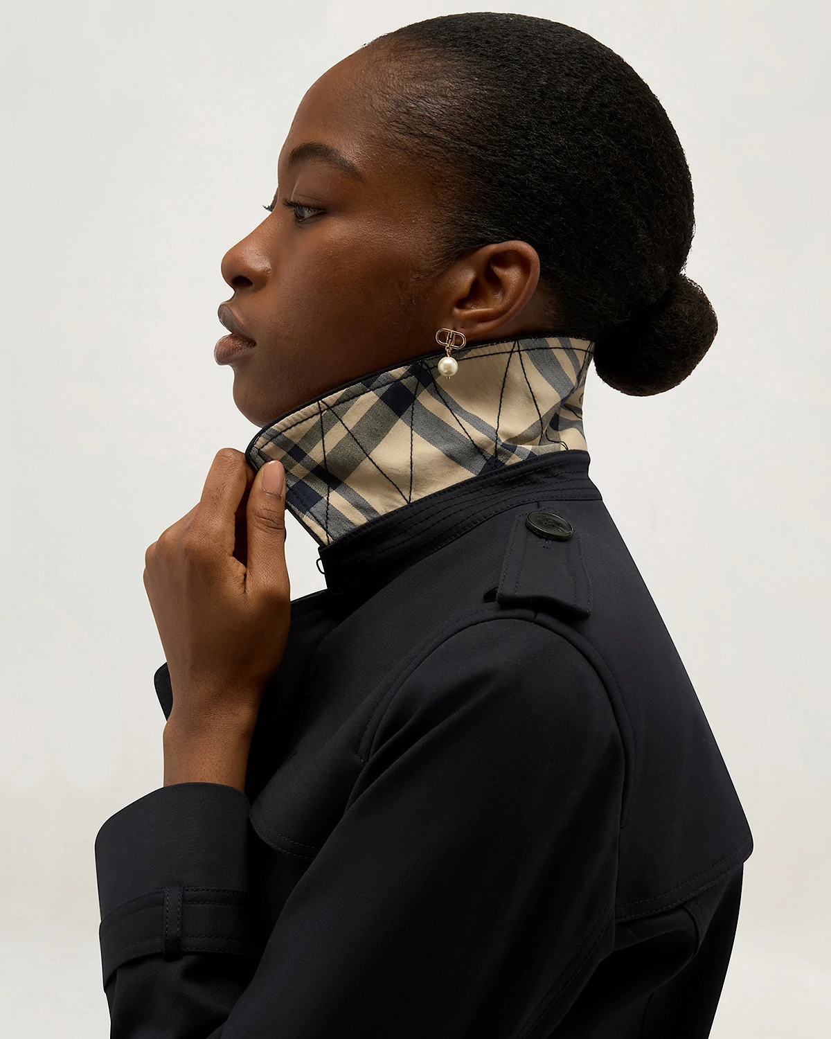 Profile of woman with dark hair in a bun wearing a black jacket with raised beige plaid collar and pearl earring.