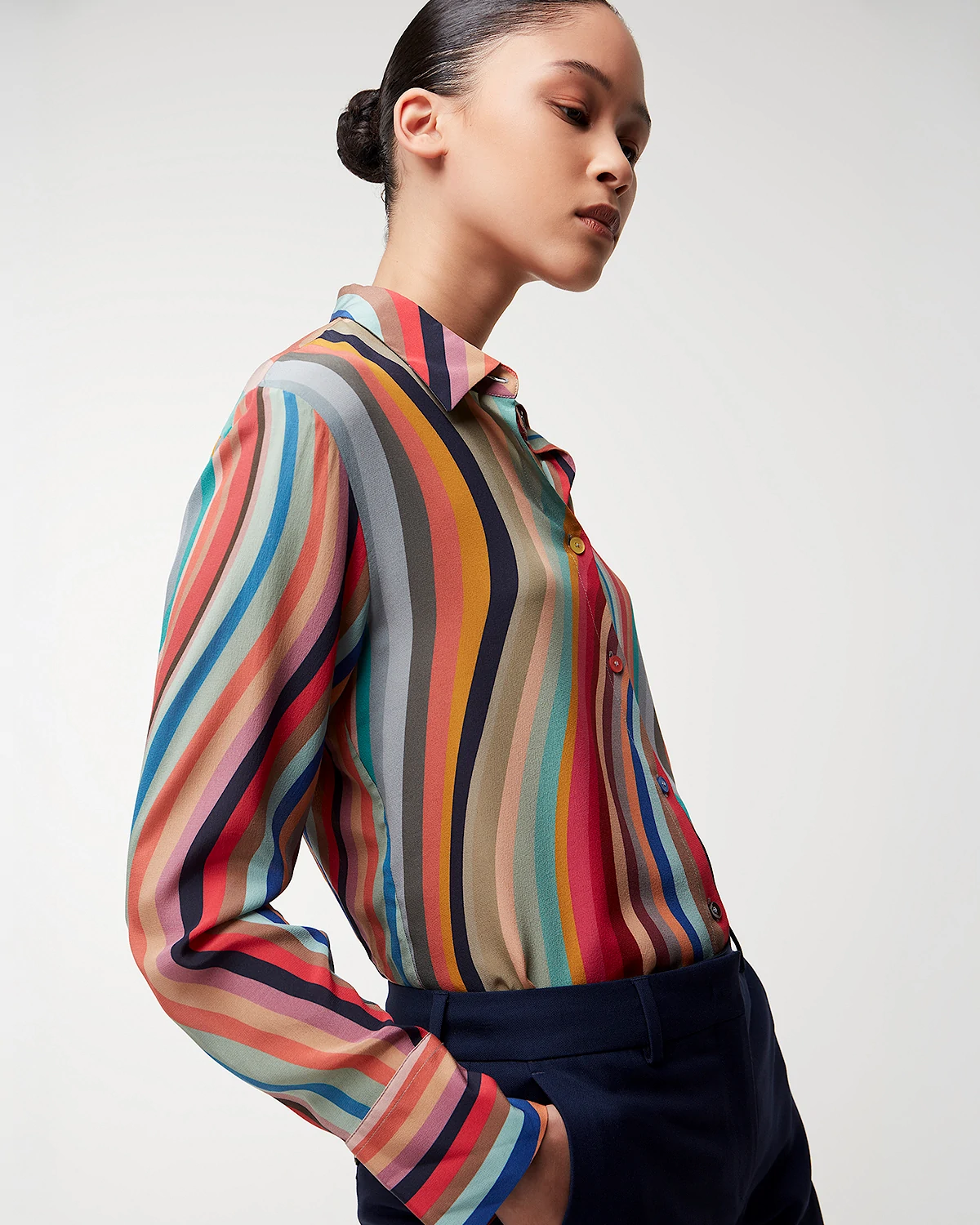Woman in a colorful vertical striped button-up Paul Smith shirt and dark pants with hand in pocket on white background.