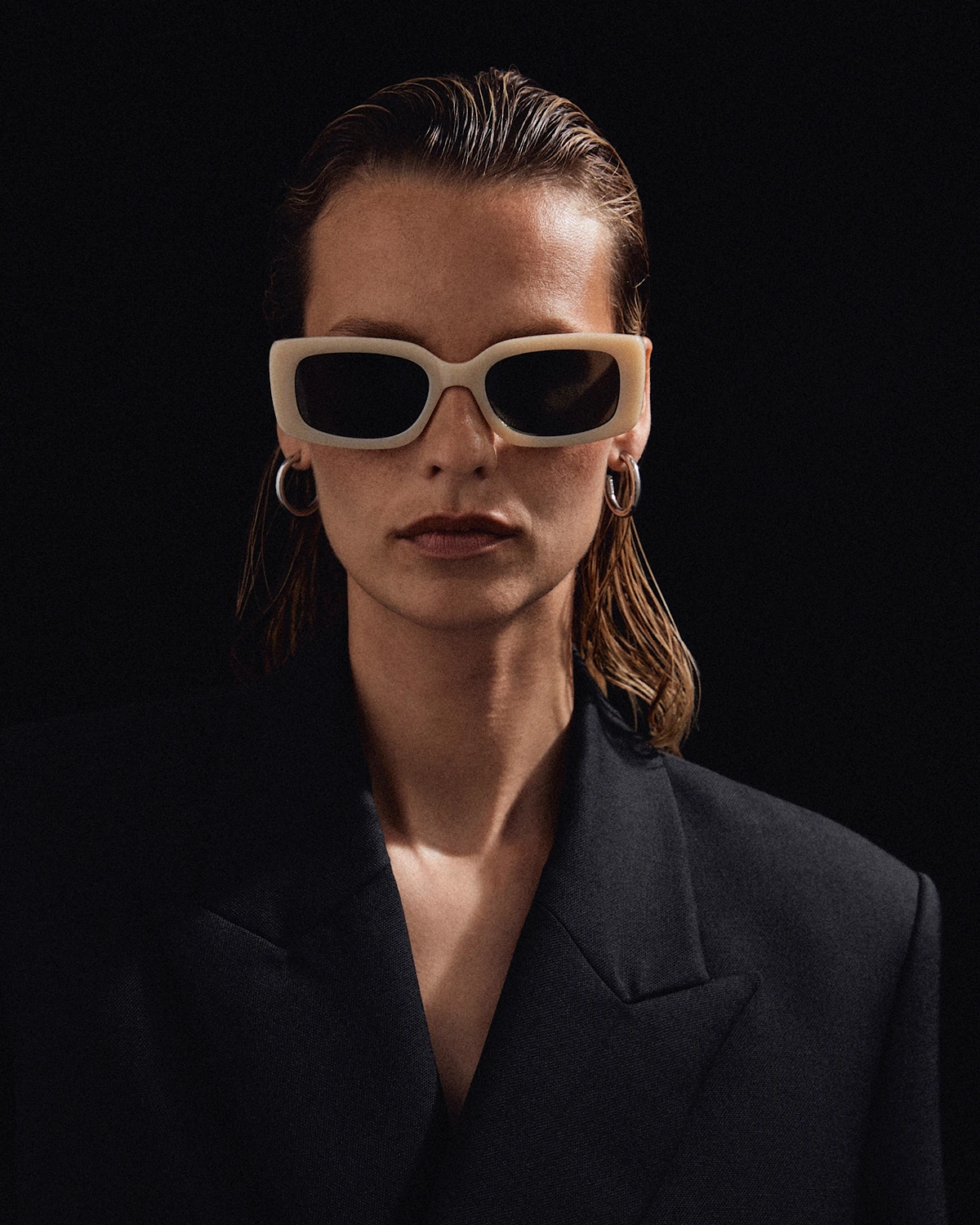 LRP fashion model with slicked-back hair wearing oversized rectangular sunglasses, hoop earrings, and a black blazer.