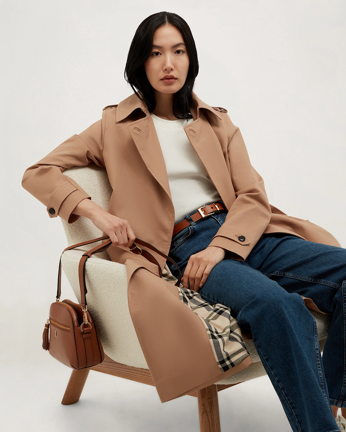 Woman with black hair in tan trench coat, white top, jeans, sitting on a white armchair holding a brown handbag.