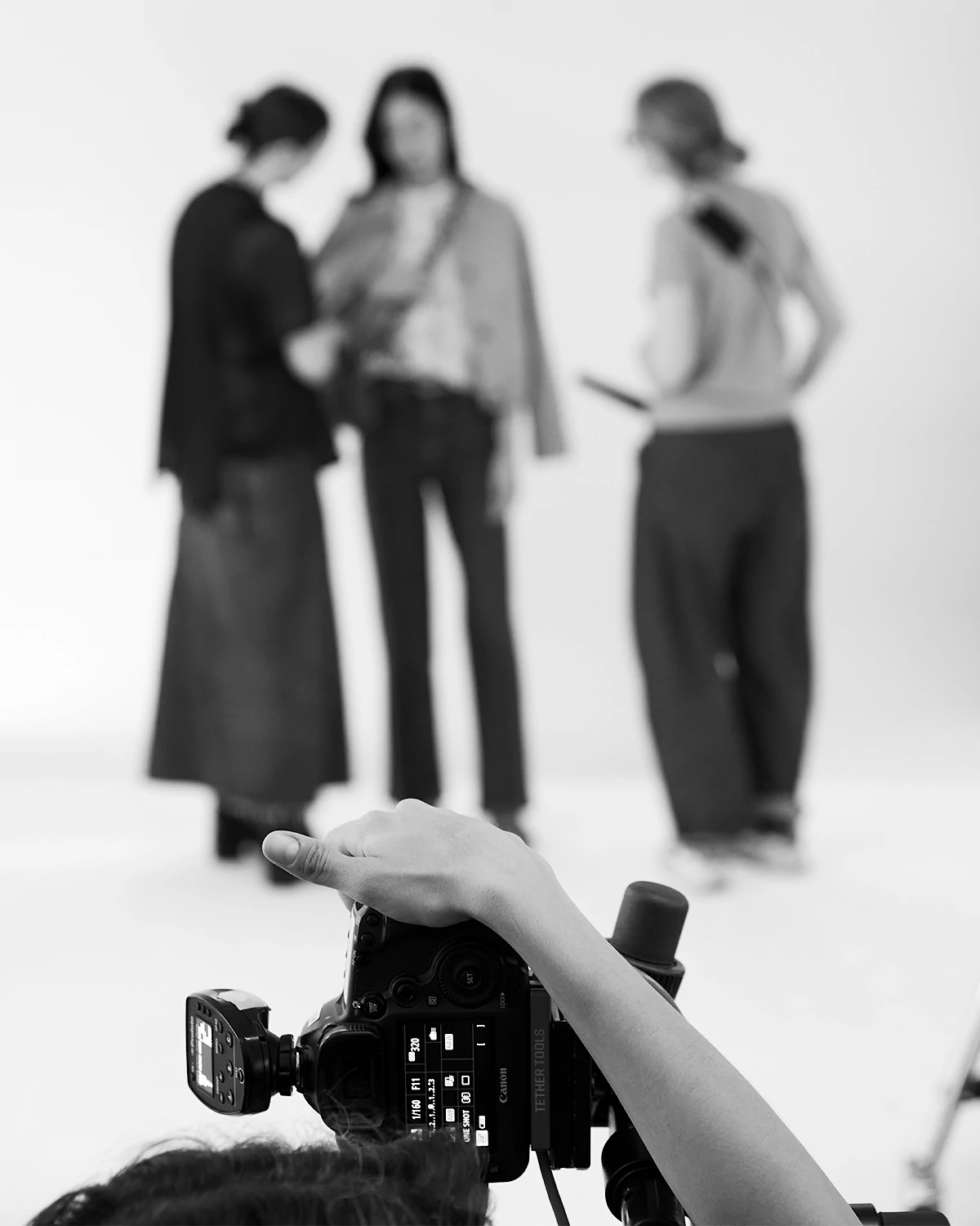 Photographer adjusting a camera, focused on three blurred people standing and talking in the background.