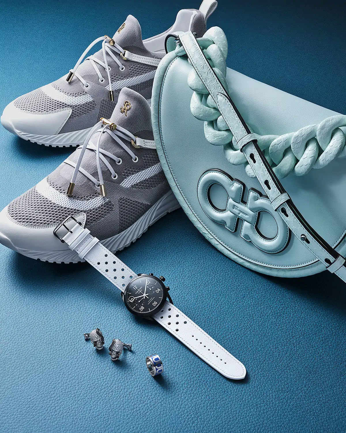 Creative Product photography of Grey sneakers, light blue handbag, white watch, and silver jewellery on a blue textured surface.