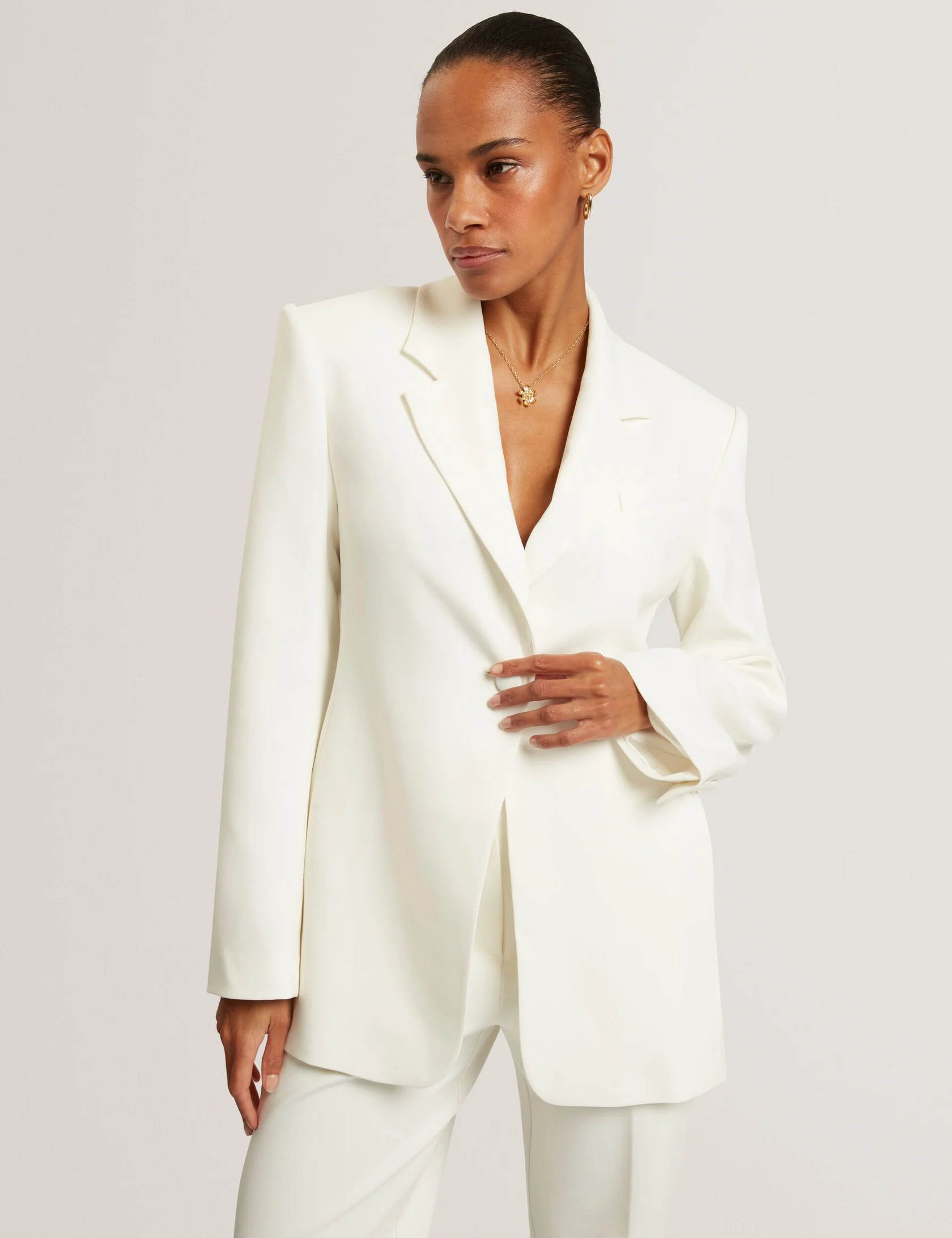 Woman wearing a white suit with a blazer and matching trousers, accessorized with gold jewelry.