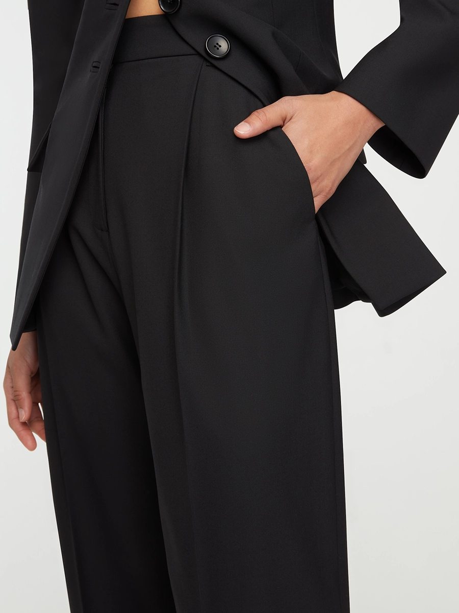 Close-up of a person wearing black Emilia Wickstead tailored pants and blazer with hand in pocket.