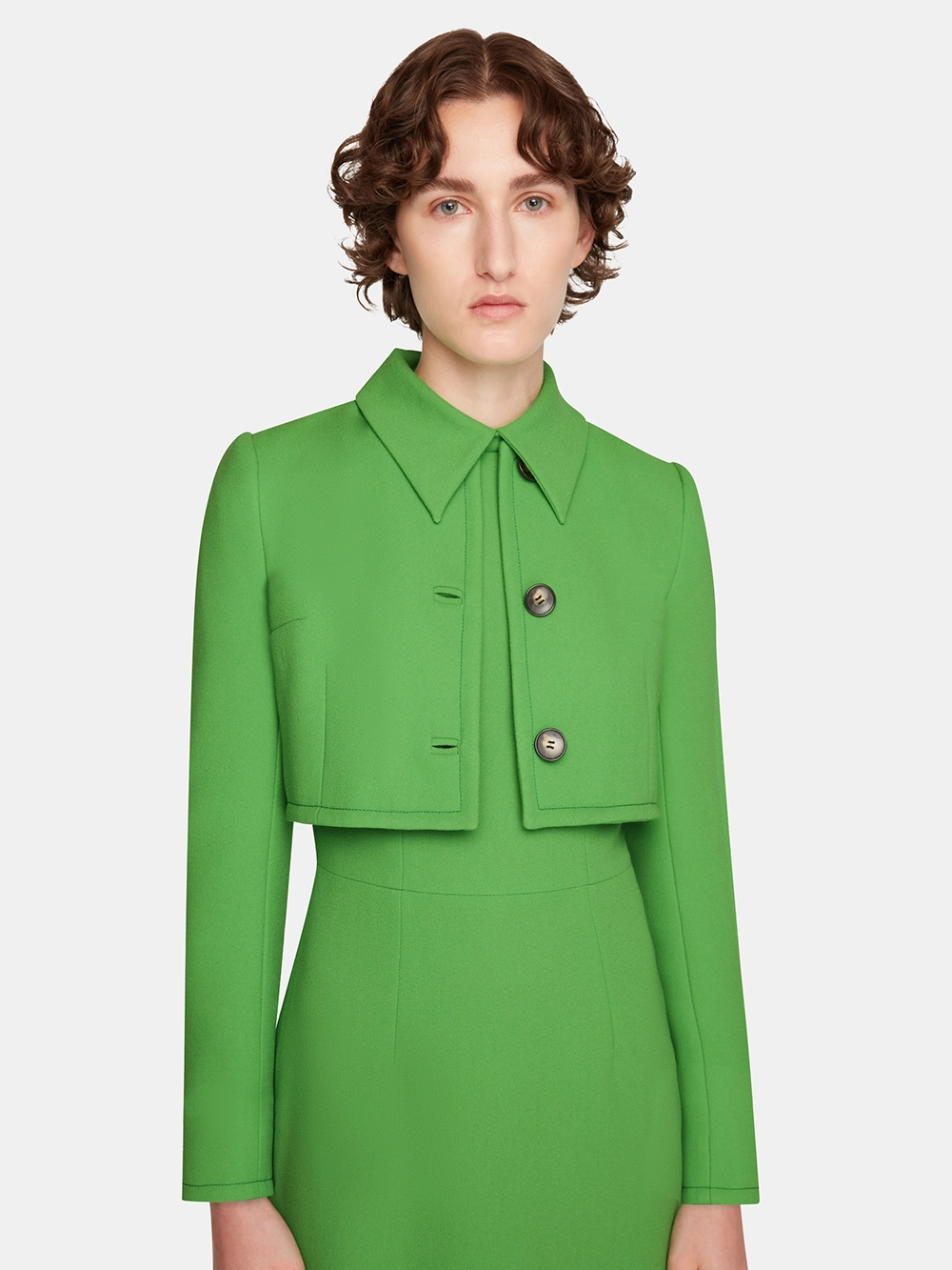 Woman with short curly hair wearing a bright green Emilia Wickstead cropped jacket over a matching green dress.