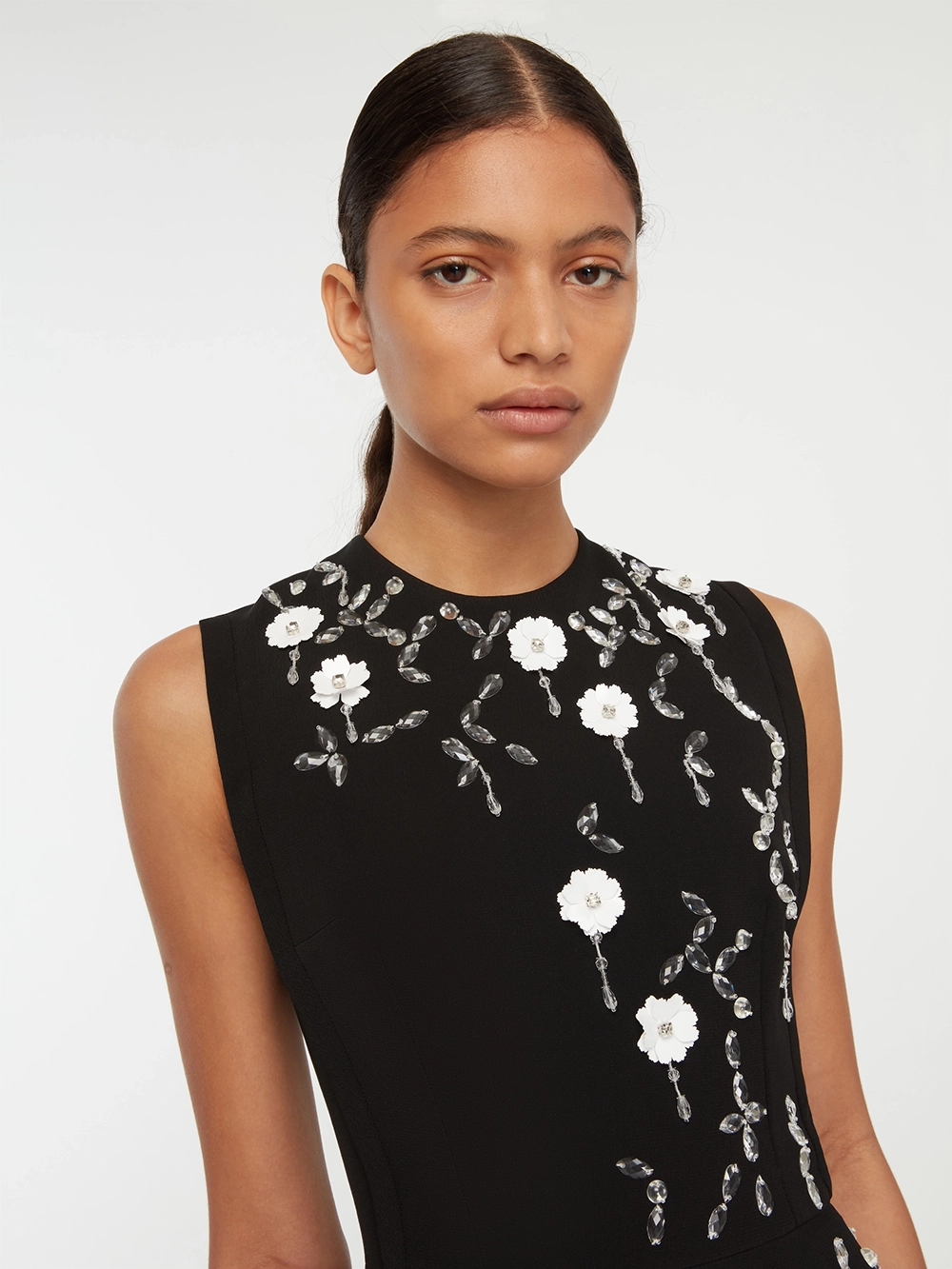 Woman wearing a sleeveless black Emilia Wickstead dress with white floral and crystal embellishments on the front.