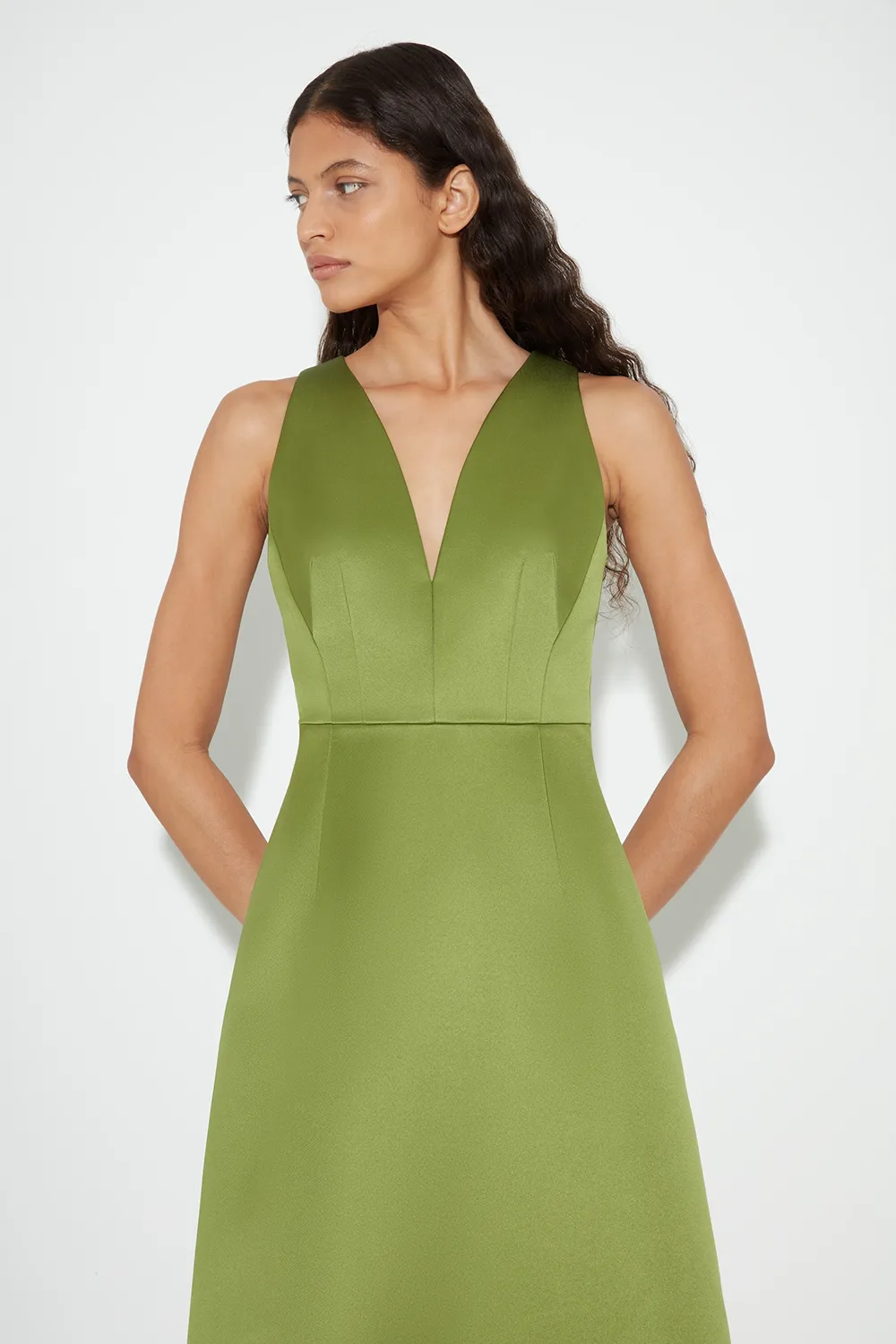 Woman with long wavy black hair wearing a sleeveless olive green Emilia Wickstead dress with deep V neckline.