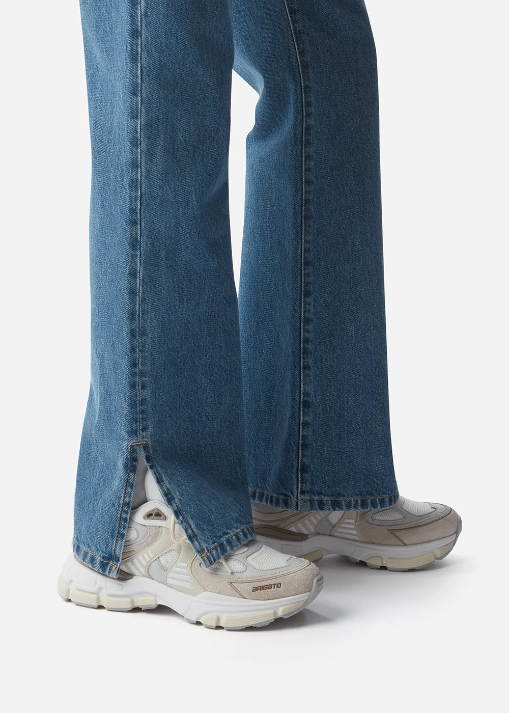 White and beige Axel Arigato sneakers with 'ARIGATO' text worn with blue denim jeans featuring side slits.