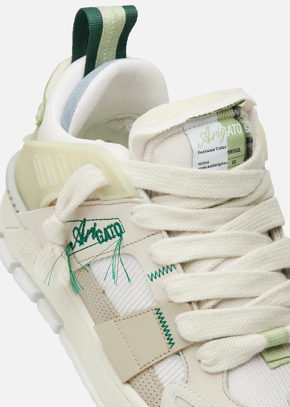 Close-up of a cream Axel Arigato sneaker with green stitching, thick cream laces, and a striped heel pull tab.