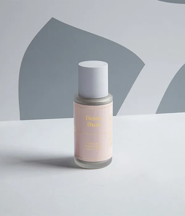 Small bottle with white cap labeled 'Detox Dust' placed on a white surface with gray abstract background.