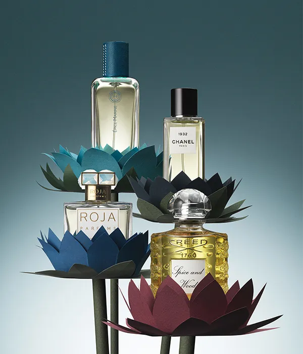 Four luxury perfume bottles displayed on stylized flower stands with dark green, blue, and maroon petals.