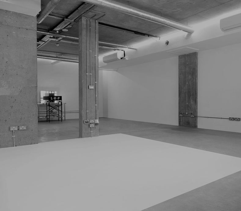 Black and white photograph of an LRP industrial studio space featuring exposed concrete pillars, a large white square on the floor, and a lighted makeup station in the background.