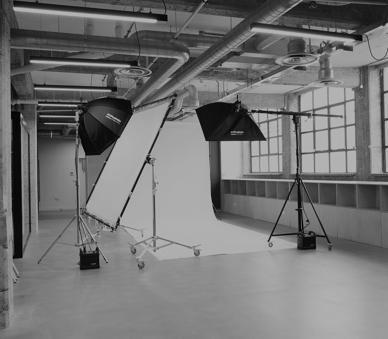 Black and white photograph of a professional LRP photo studio setup featuring a large white seamless paper backdrop, Profoto softboxes on heavy-duty stands, and a large rectangular diffusion panel inside an industrial-style room with exposed ceiling pipes.