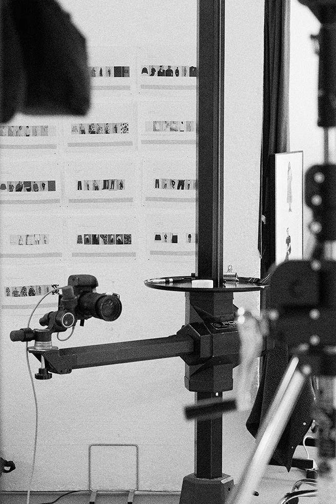 Black and white image of a professional camera mounted on a heavy studio stand at LRP studios, with a detailed printed shot list on the wall behind it, prepared for eCommerce photography.