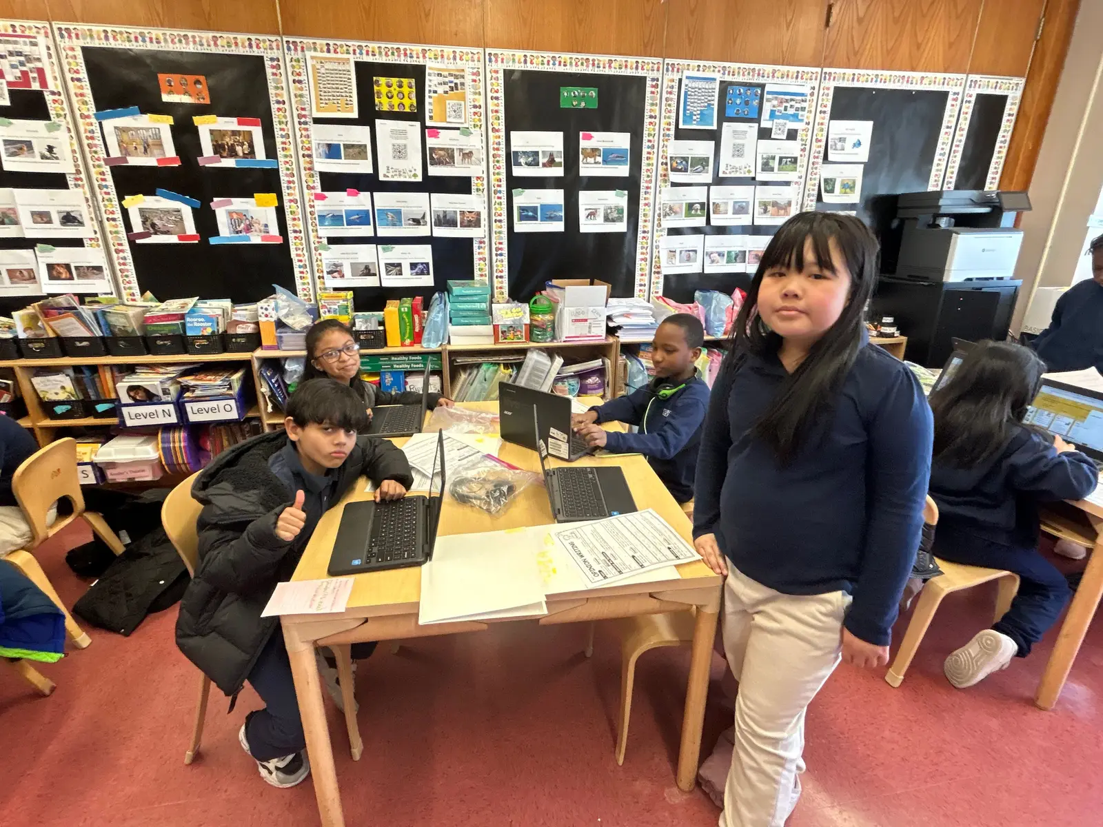 Students working on laptops at classroom table with bulletin boards