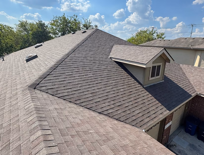 Gray shingled residential roof with a small dormer under blue sky and scattered clouds.
