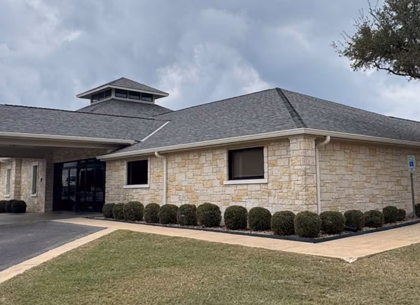 Stone building with a gray roof, bushes lining the foundation, and a paved walkway under a cloudy sky.