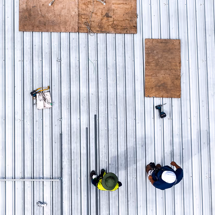 Overhead view of two construction workers on a metal roof with tools and wooden boards nearby.