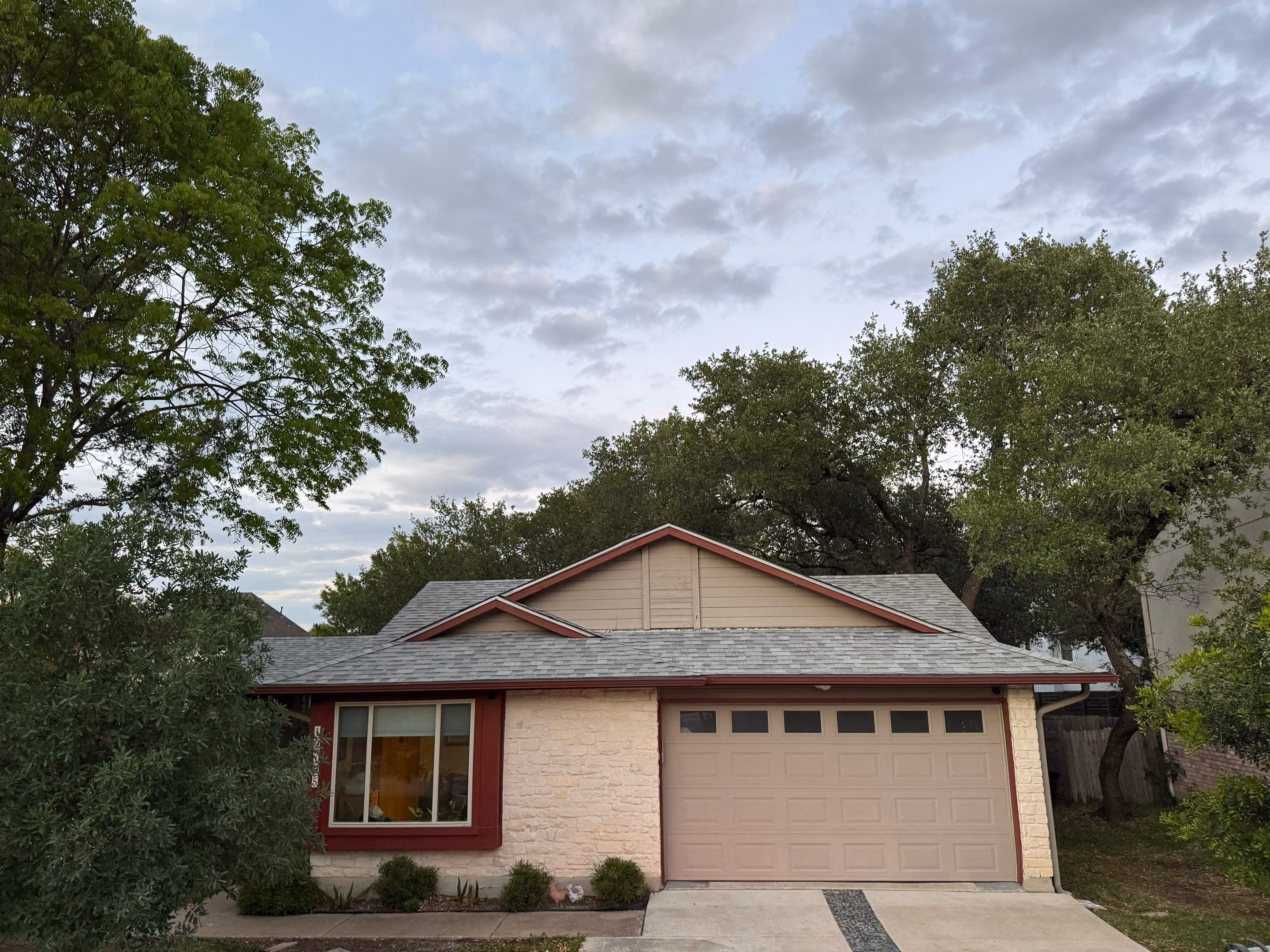 Single-story house with red-trimmed window and beige garage door surrounded by green trees.