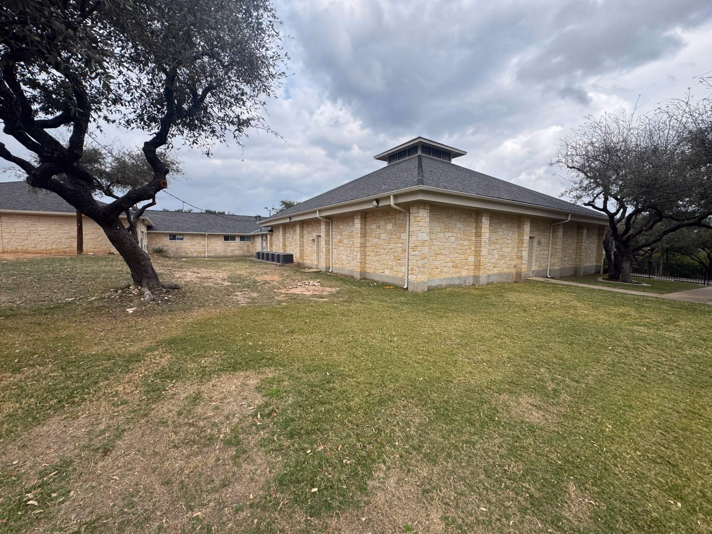 Stone building with a hip roof and stone walls in a grassy yard with two large trees under cloudy sky.