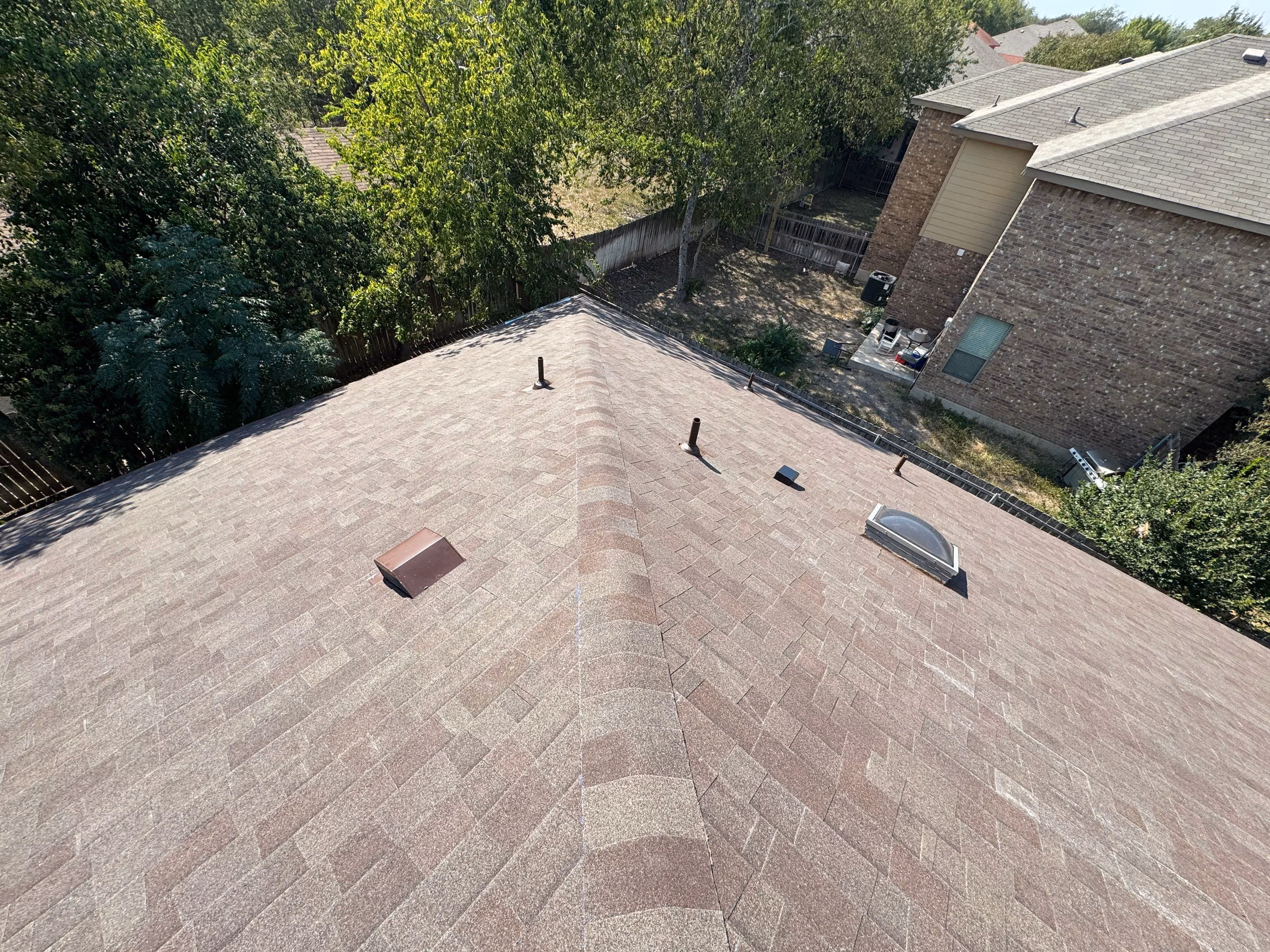 Brown shingled roof with vents, a skylight, and nearby trees and brick houses.