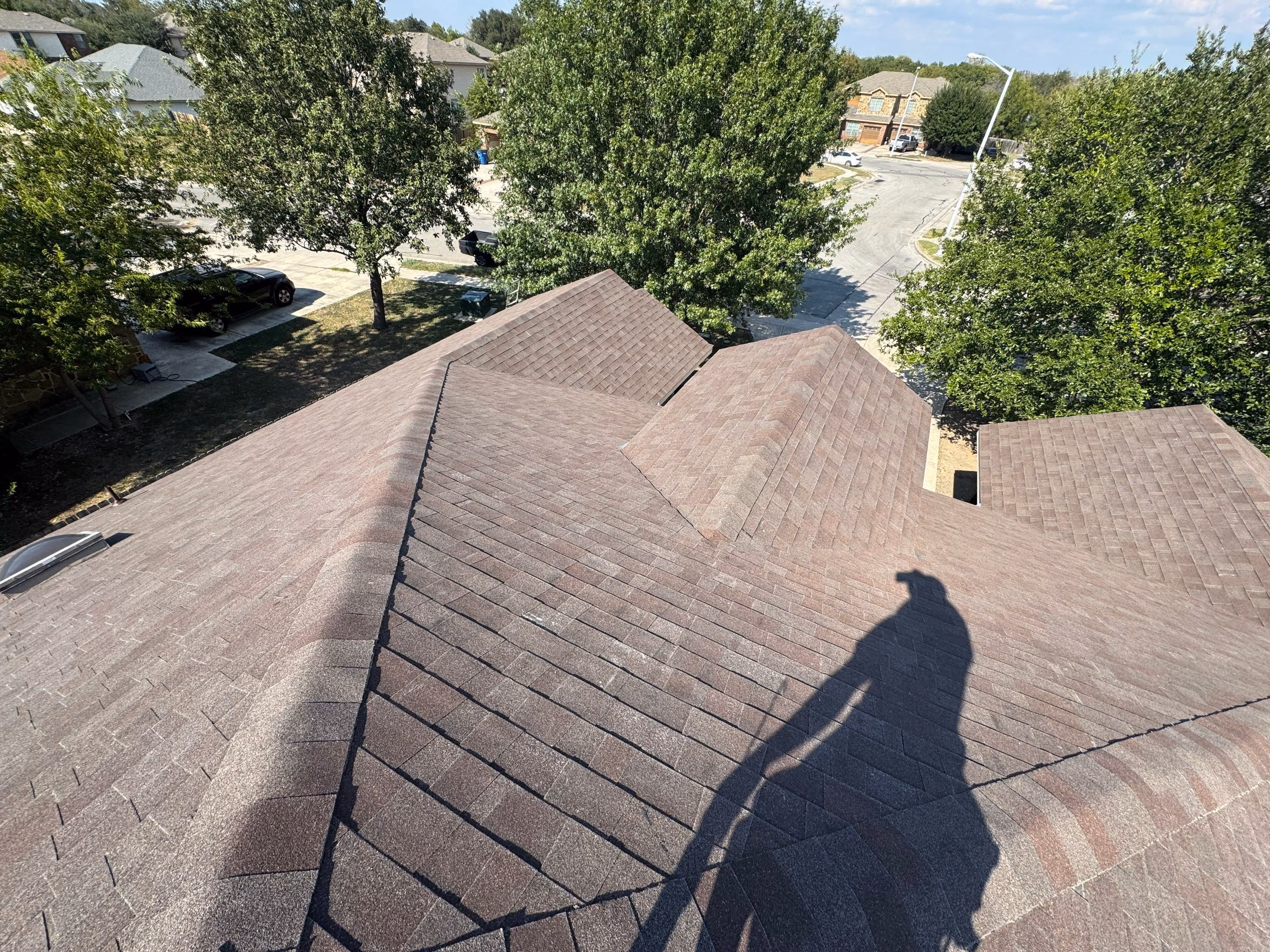 Shadow of a person on brown shingled roof with trees and houses in the background.