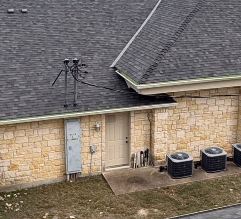 Stone building wall with beige door, electrical box, three air conditioning units, and black roof shingles.